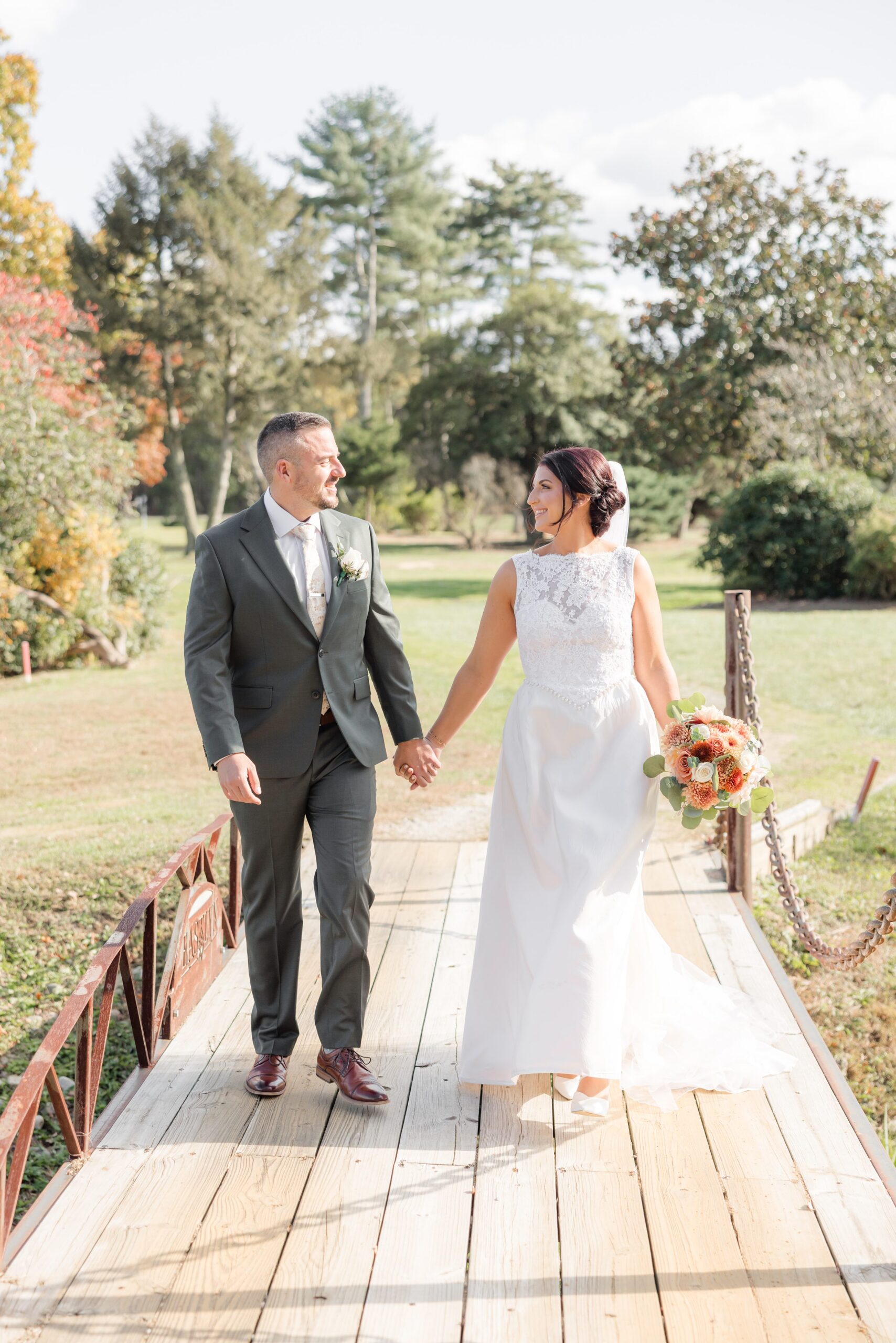 Bride and groom walk across a sunlit bridge, gazing at each other with quiet joy as soft autumn colors surround the beginning of their forever