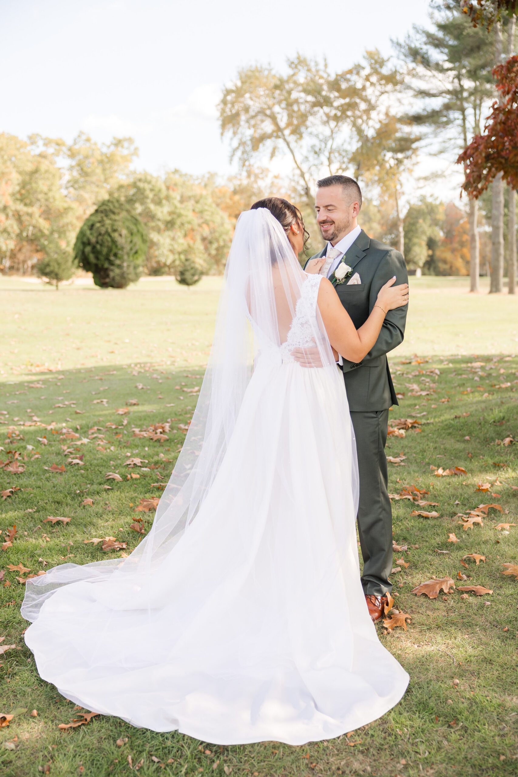 Bride and groom stand close in a sunlit field, her veil flowing softly behind her as they gaze into each other’s eyes, lost in a quiet, romantic moment together