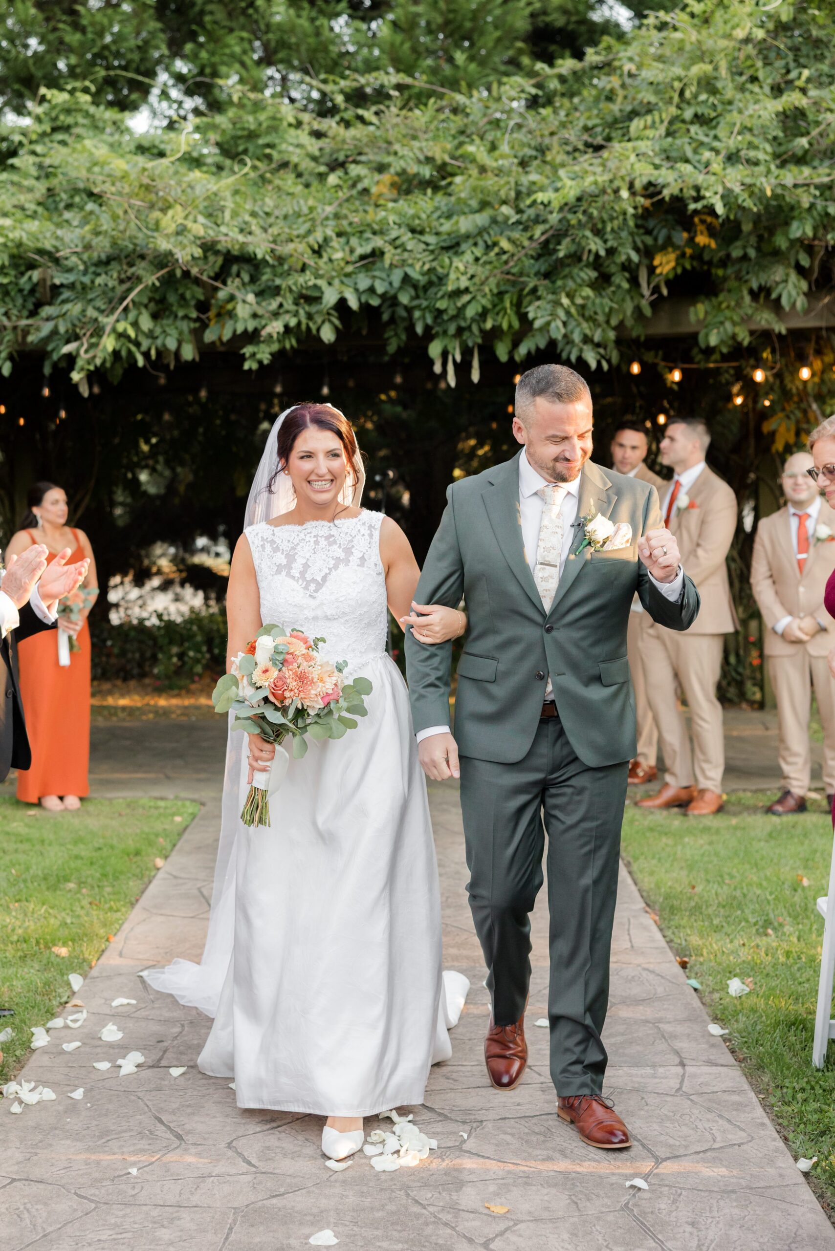 Bride and groom walk down together the aisle, smiling softly as petals scatter beneath their feet