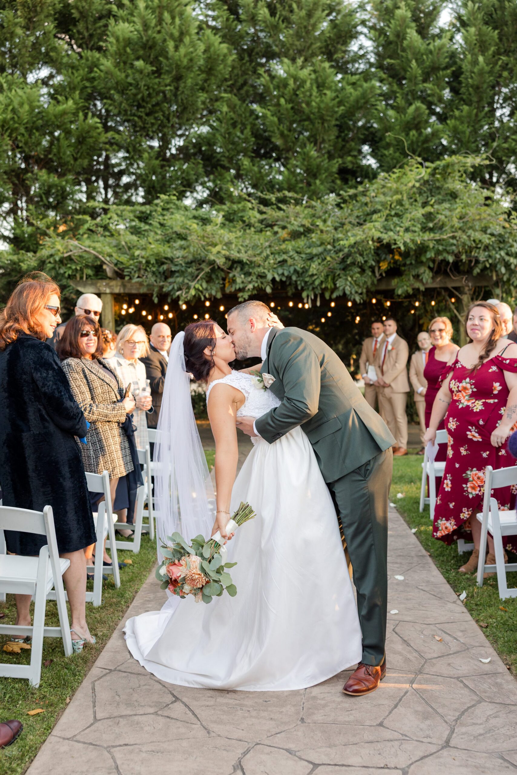 Newlywed couple shares their first kiss as husband and wife beneath a canopy of greenery and glowing lights, surrounded by loved ones