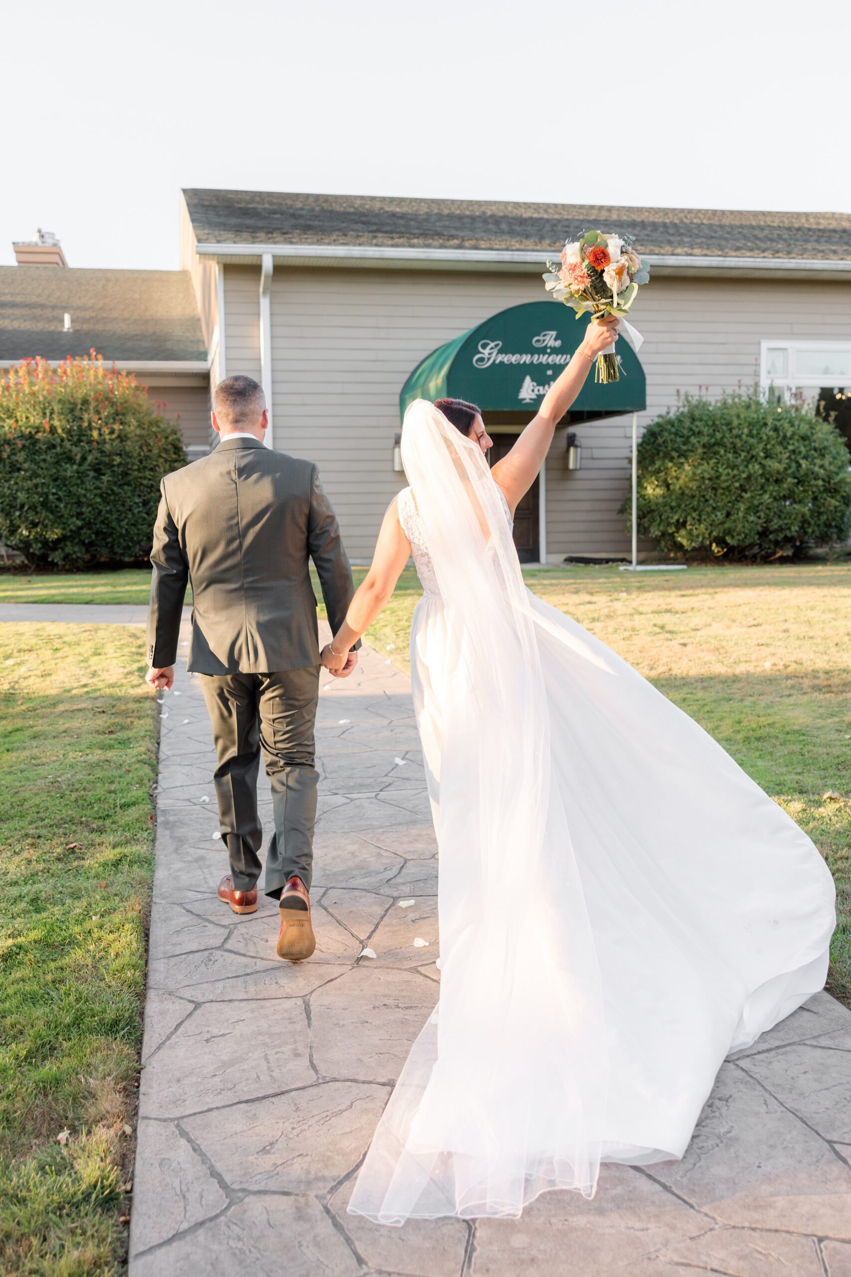 Bride and groom walk together down a sunlit path, her veil trailing behind as she lifts her bouquet in a joyful, celebratory gesture