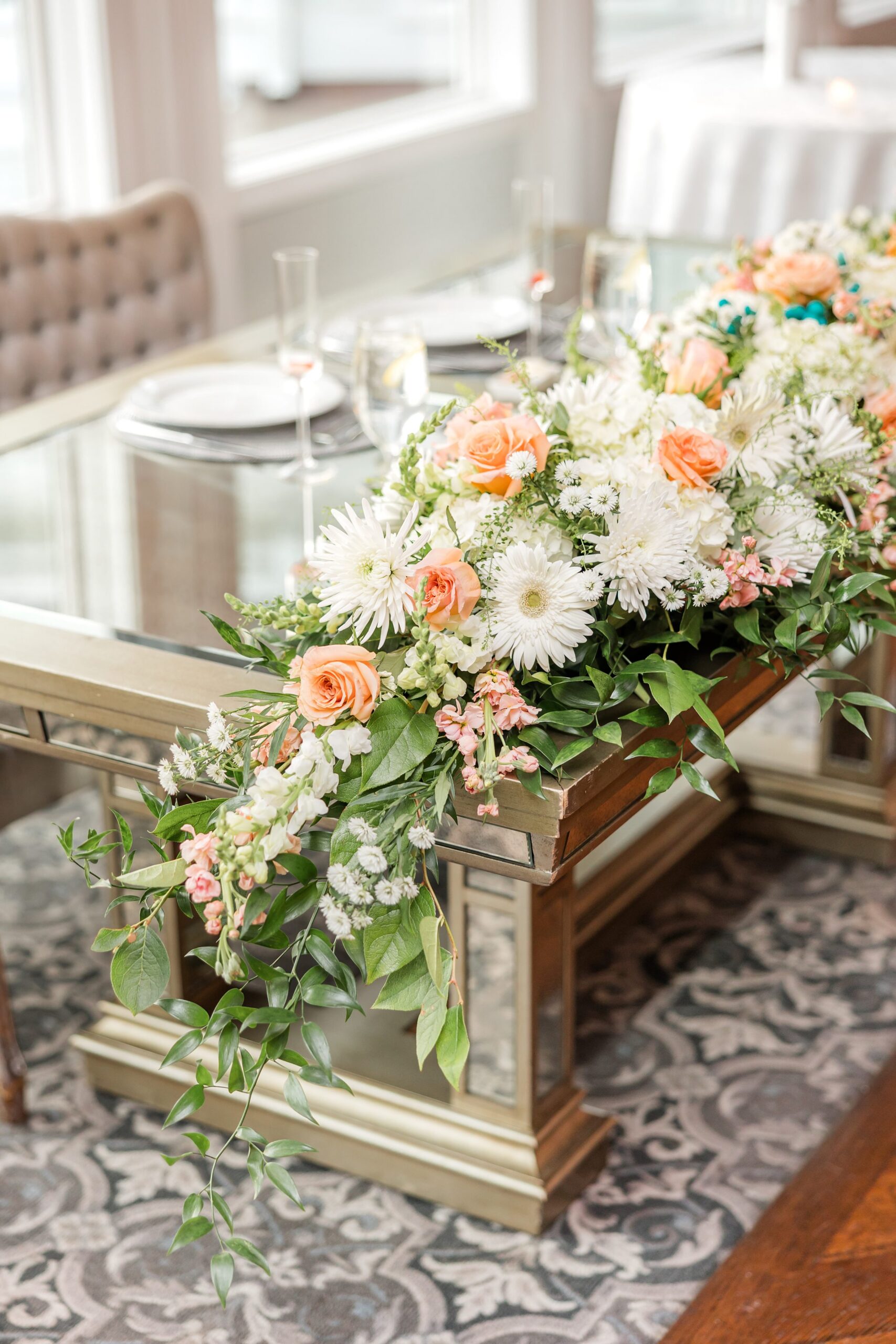 Detail shot of sweetheart table. Glass tabletop on brown frame, decorated with white and orange flowers, along with leafy greens