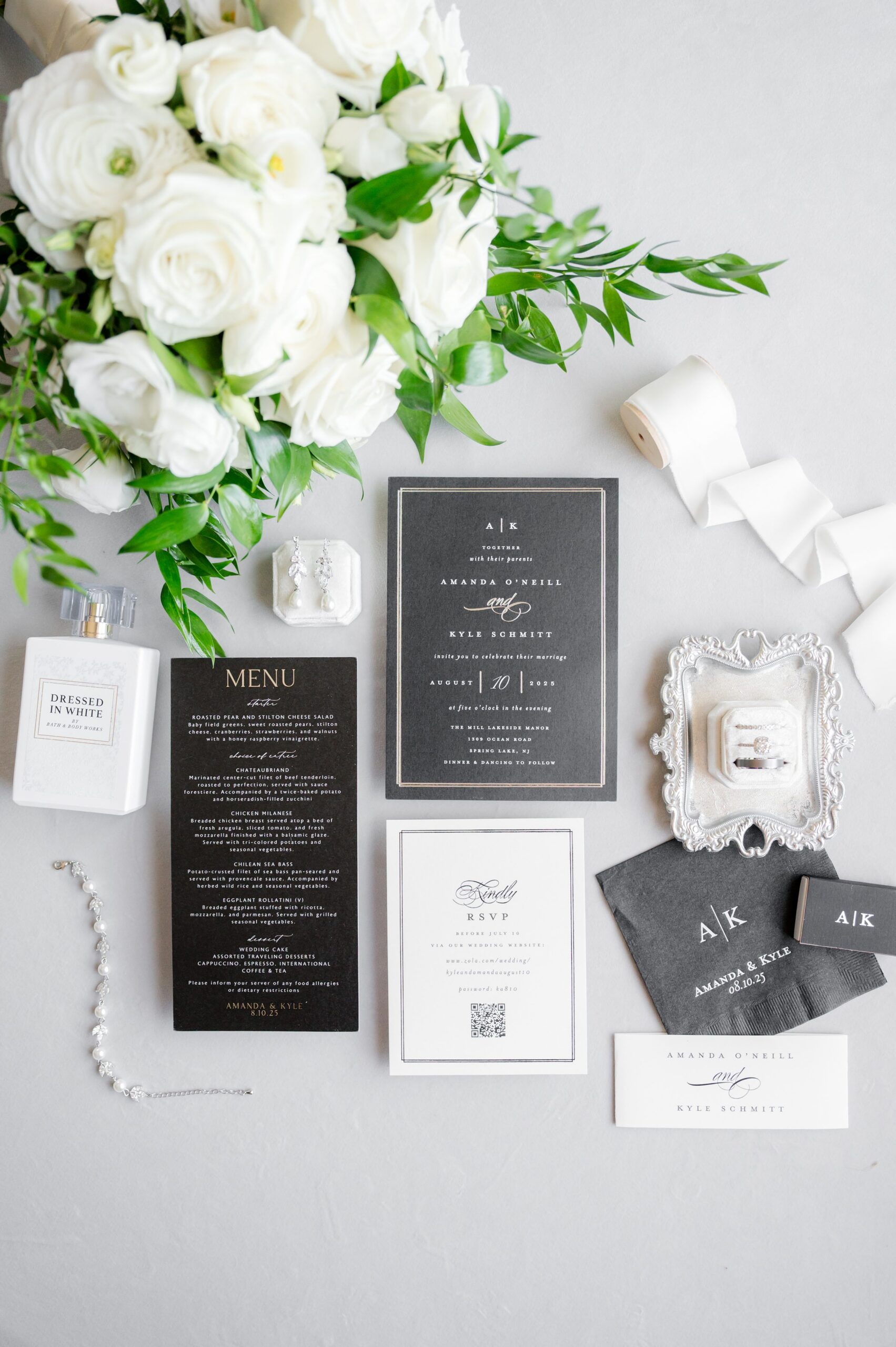 Wedding stationery flat lay with white roses and greenery on a light background, featuring black-and-white invitation suite, RSVP and menu cards, ribbon, small perfume bottle, pearl jewelry, and coordinated envelopes in a minimalist, elegant arrangement