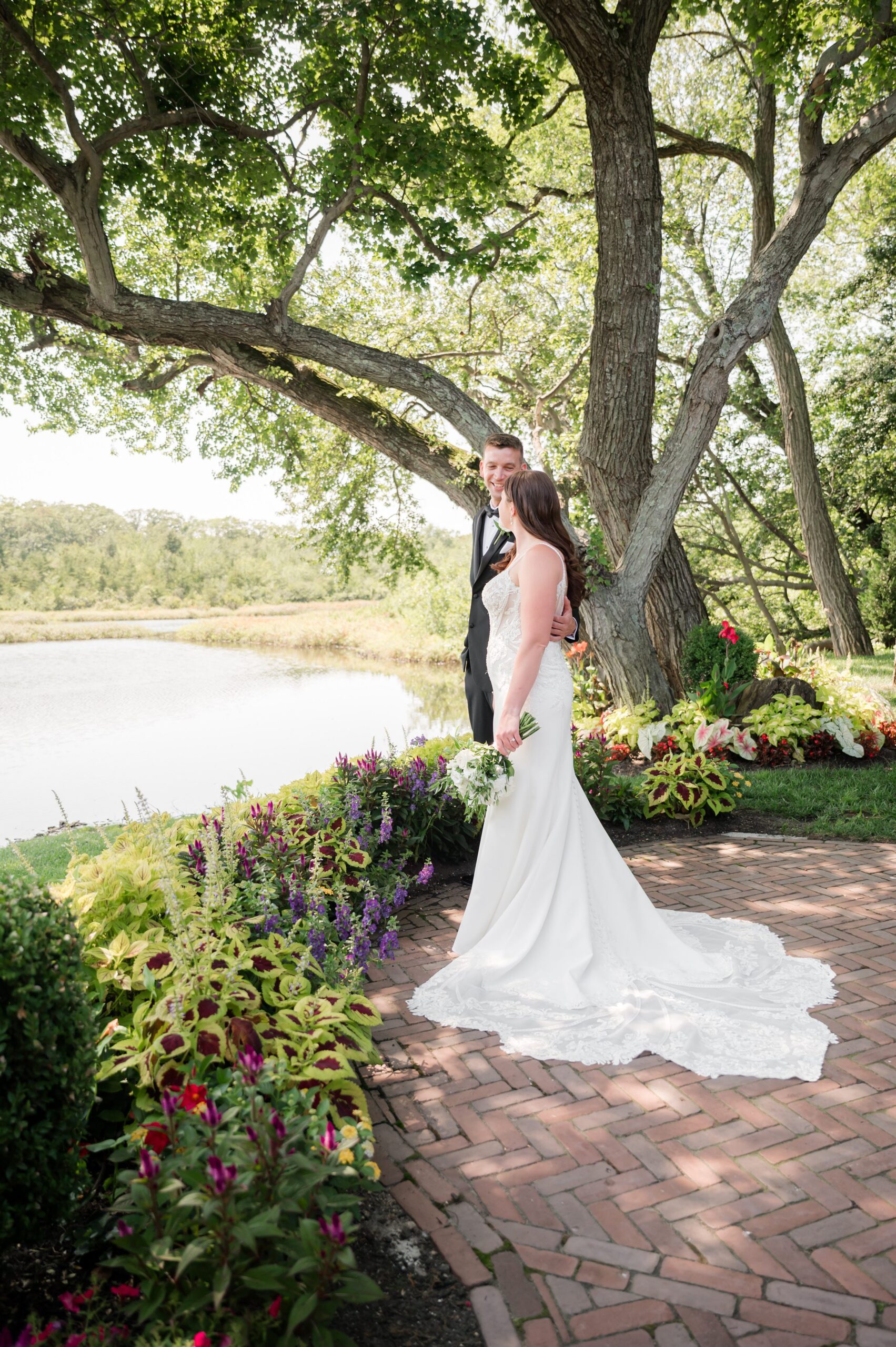 Bride and groom embracing on a garden path by the water, surrounded by lush greenery and flowers, sharing a romantic moment