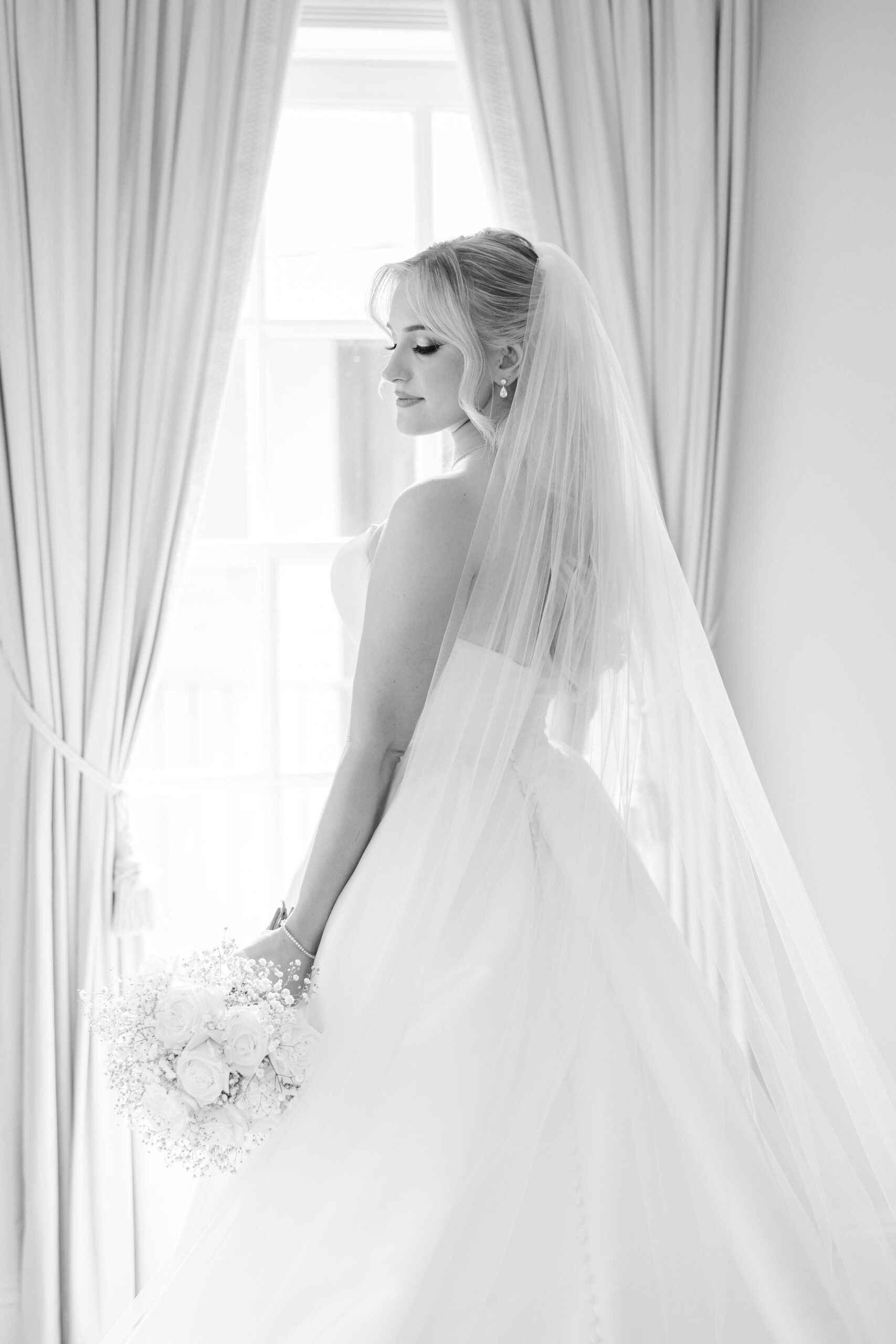 Black and white photo of a bride in her wedding dress and veil standing by a window, holding a bouquet