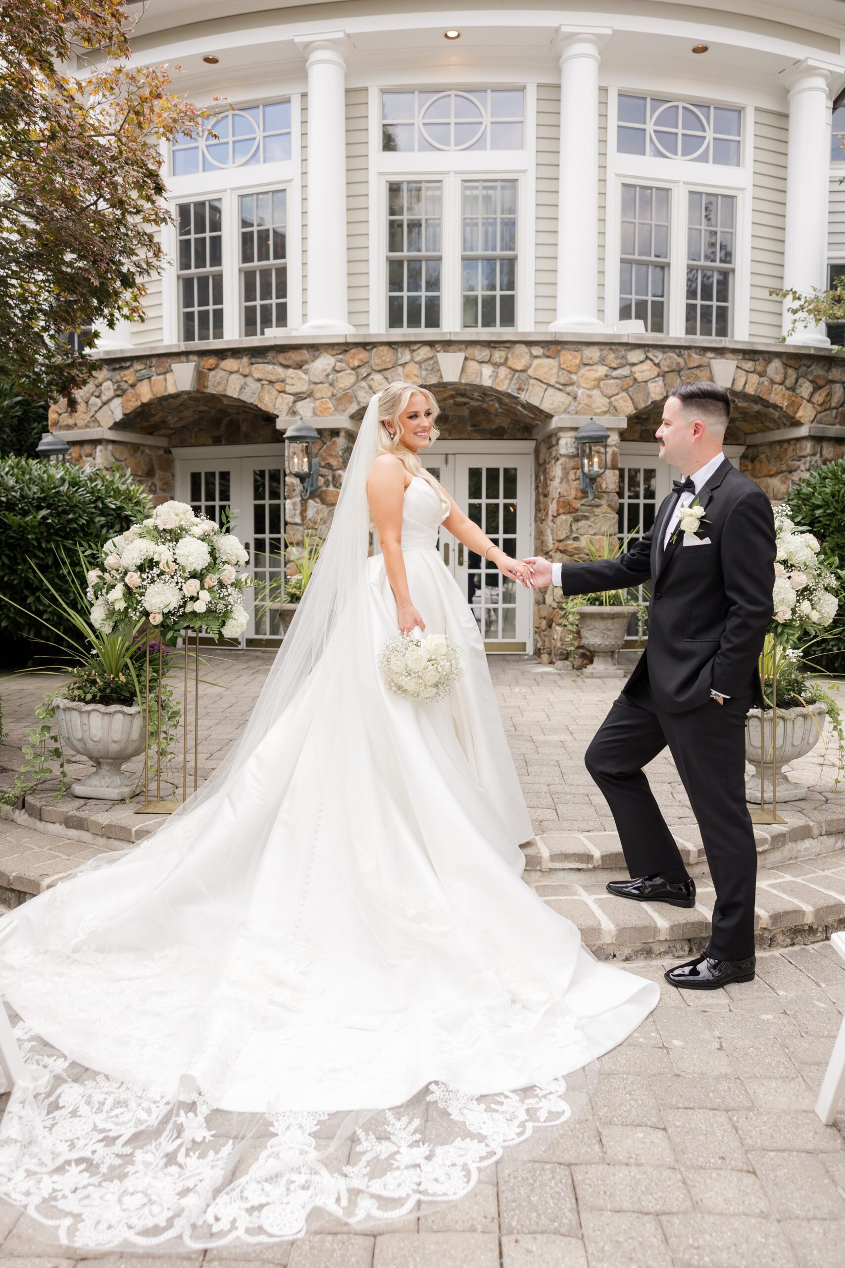 Bride and groom holding hands outdoors, with her long veil and train flowing behind her in front of a stone building