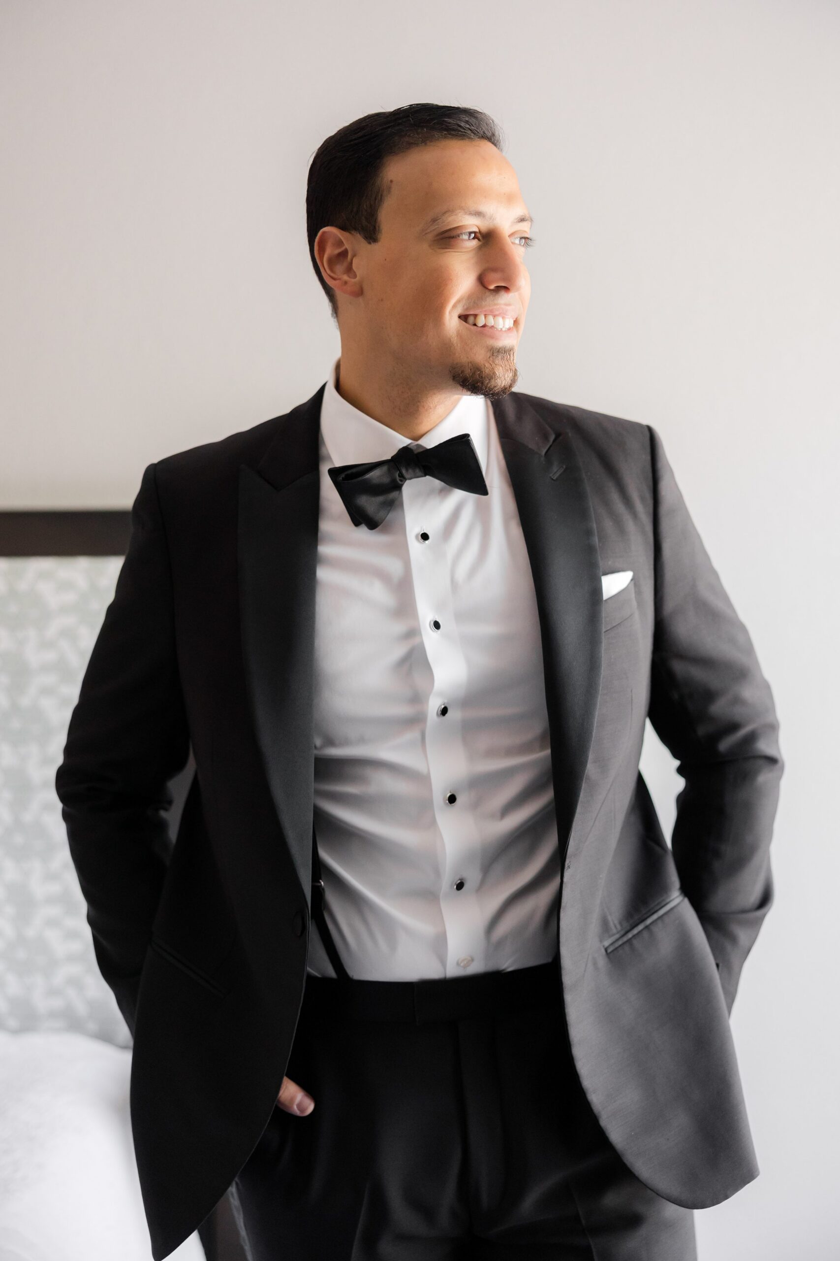 Groom in a black tuxedo standing and smiling as he looks off to the side