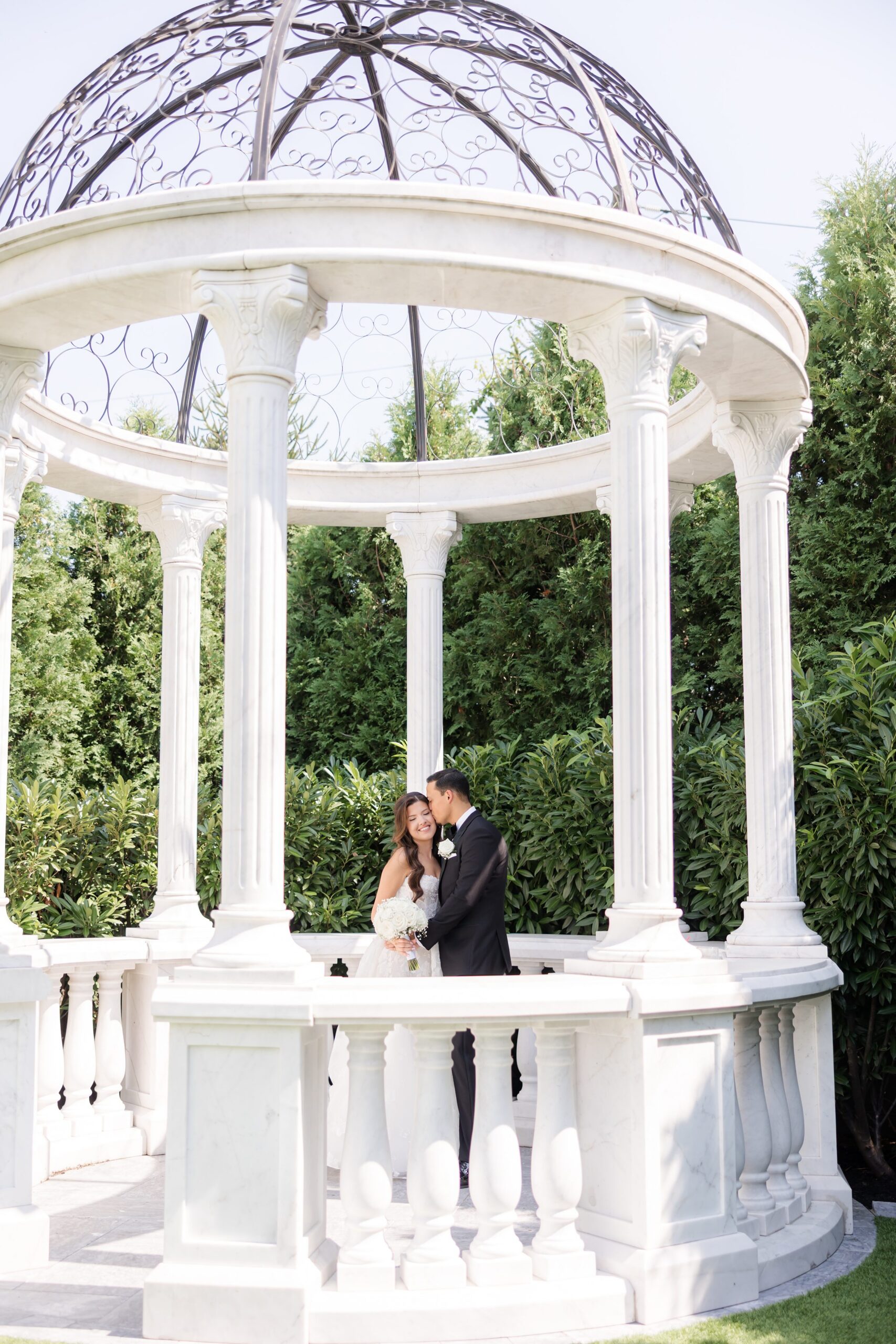 Bride and groom sharing a kiss under a white gazebo surrounded by greenery