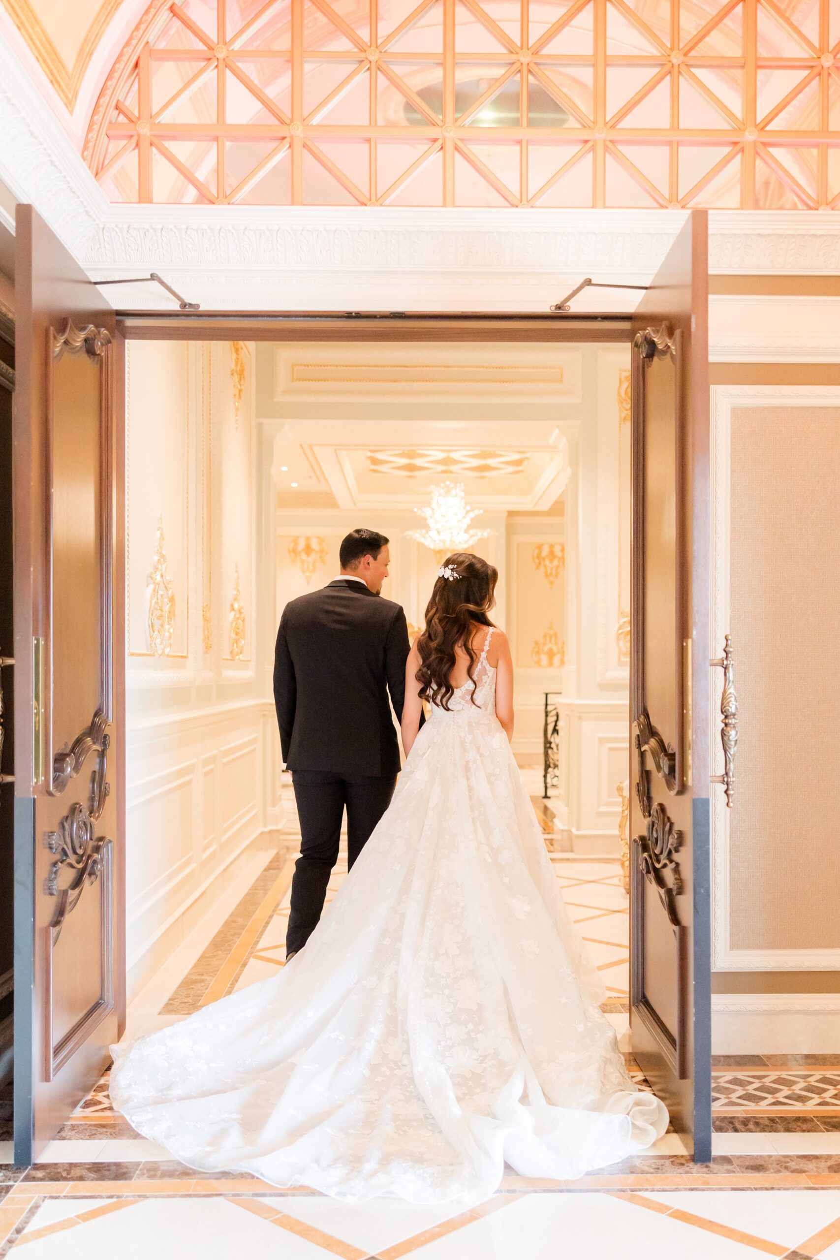 Bride and groom walk together through grand doors, her long gown trailing behind in a romantic, elegant setting