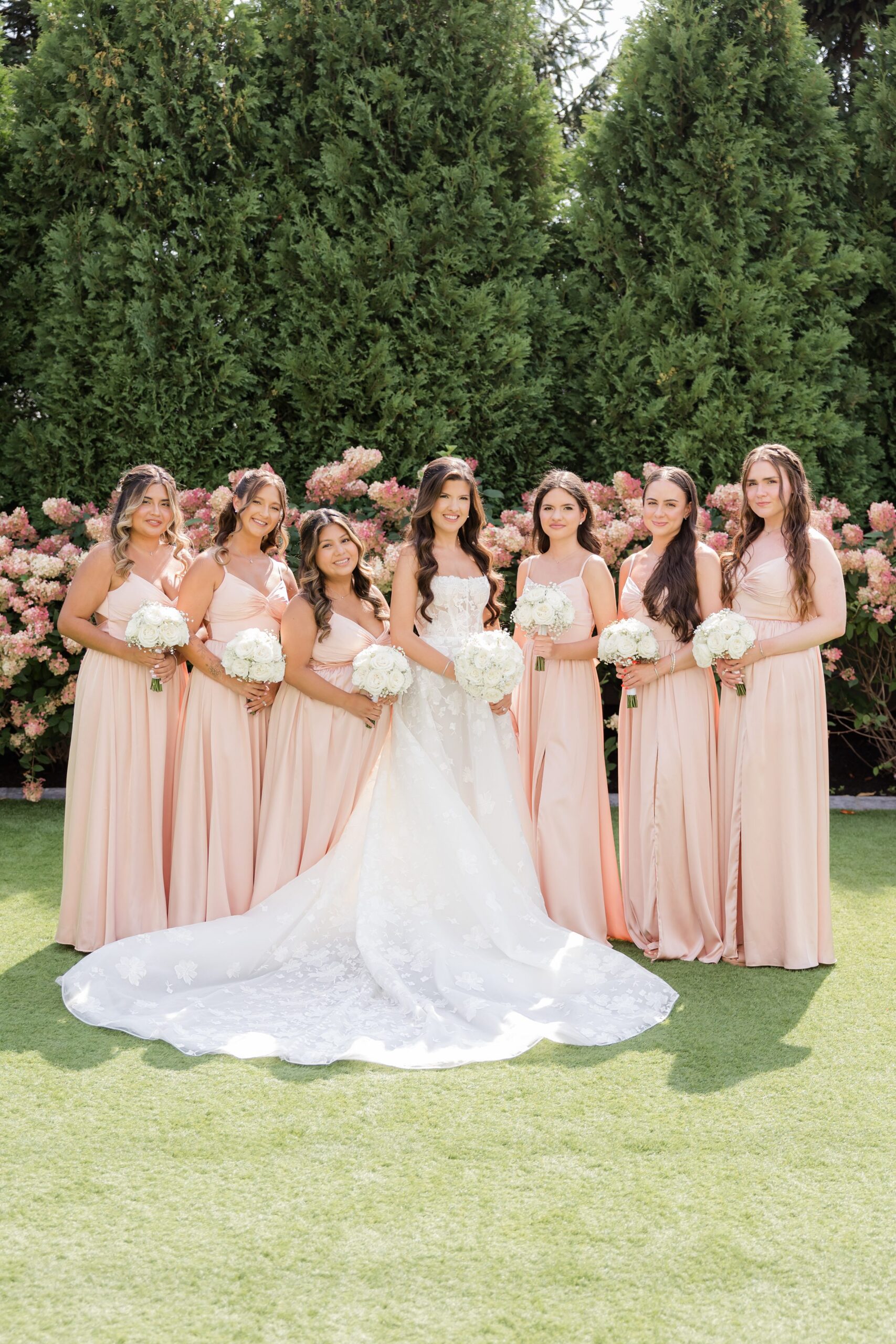 Bride stands with her bridesmaids in blush dresses, holding bouquets in a lush, romantic garden