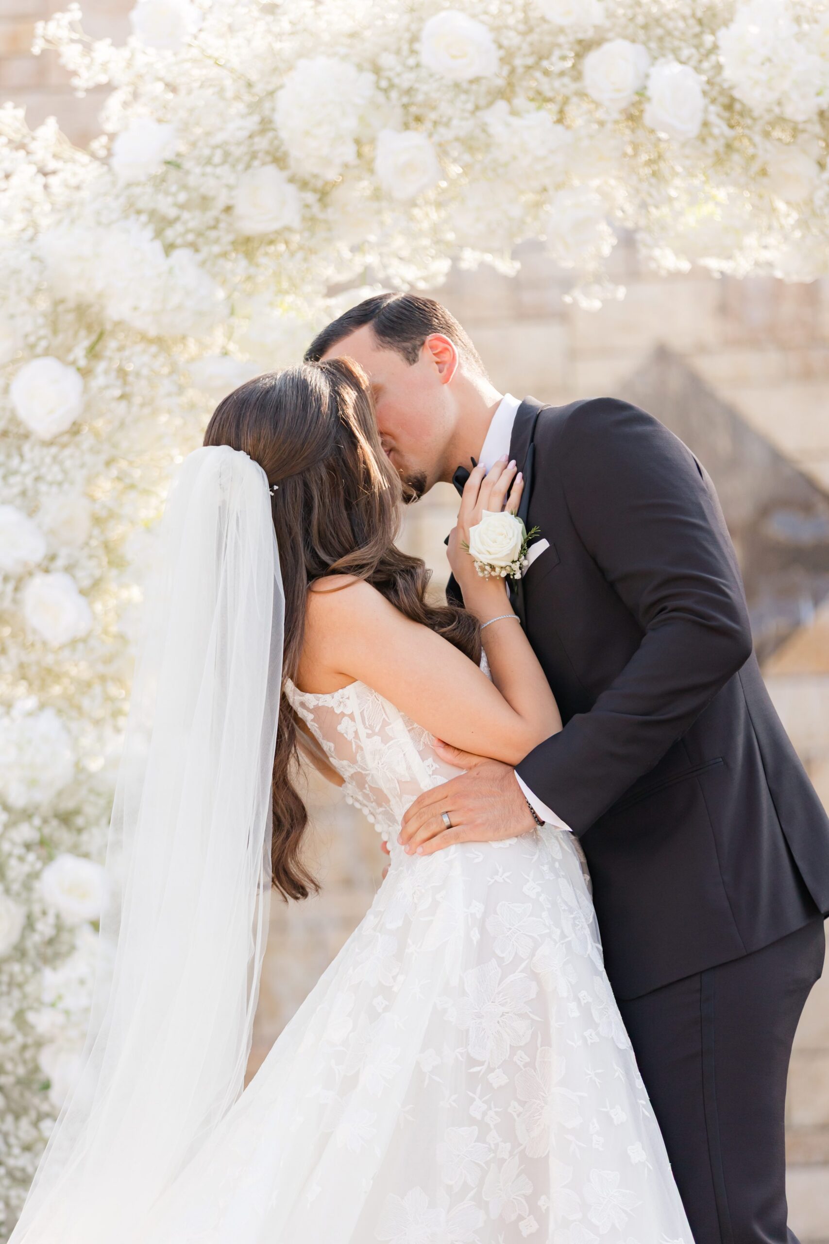 Bride and groom share a romantic kiss beneath a soft floral backdrop during their ceremony