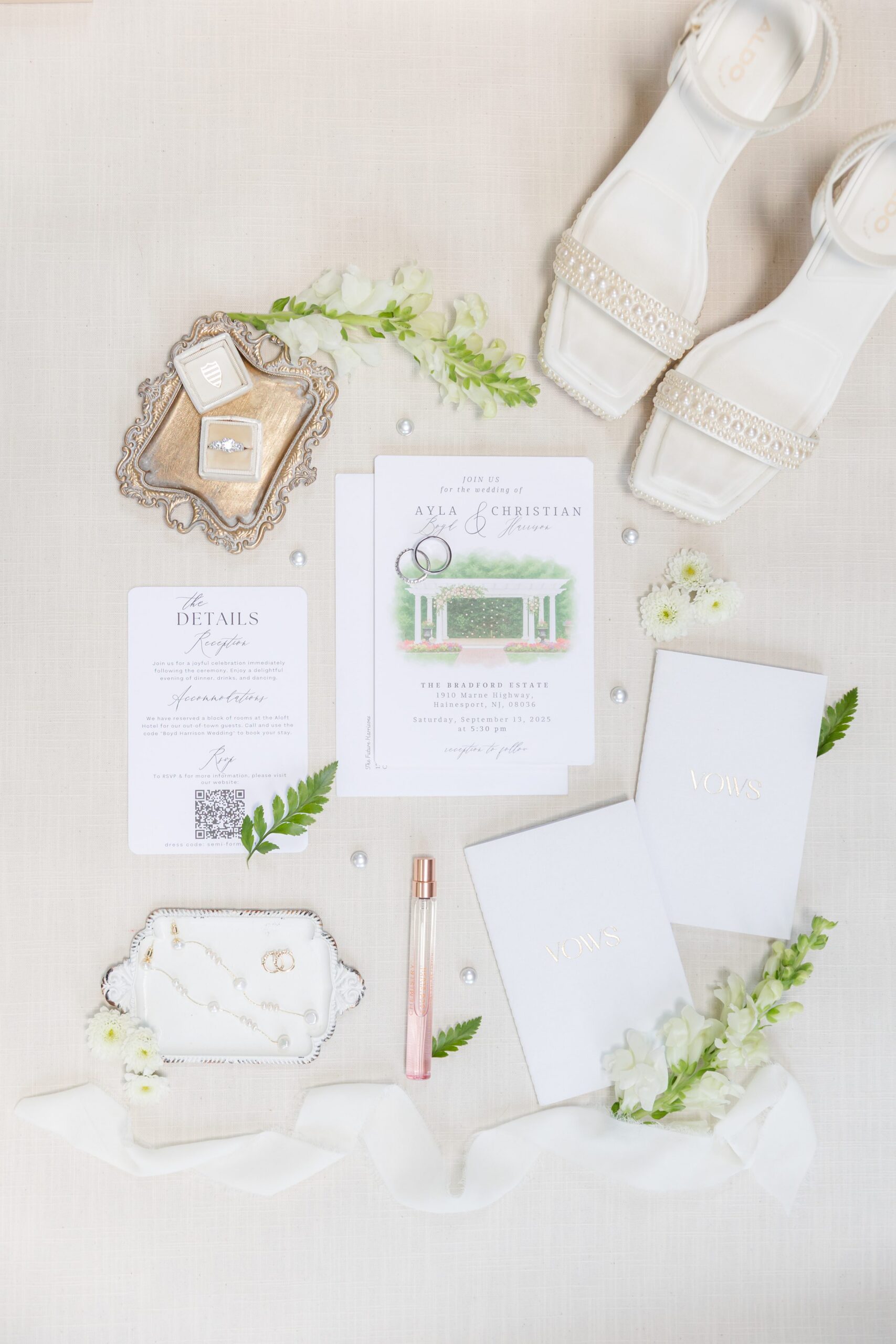 Wedding flat lay featuring invitation suite, vow books, perfume, jewelry box with engagement ring, shoes, and floral accents arranged neatly on a light surface