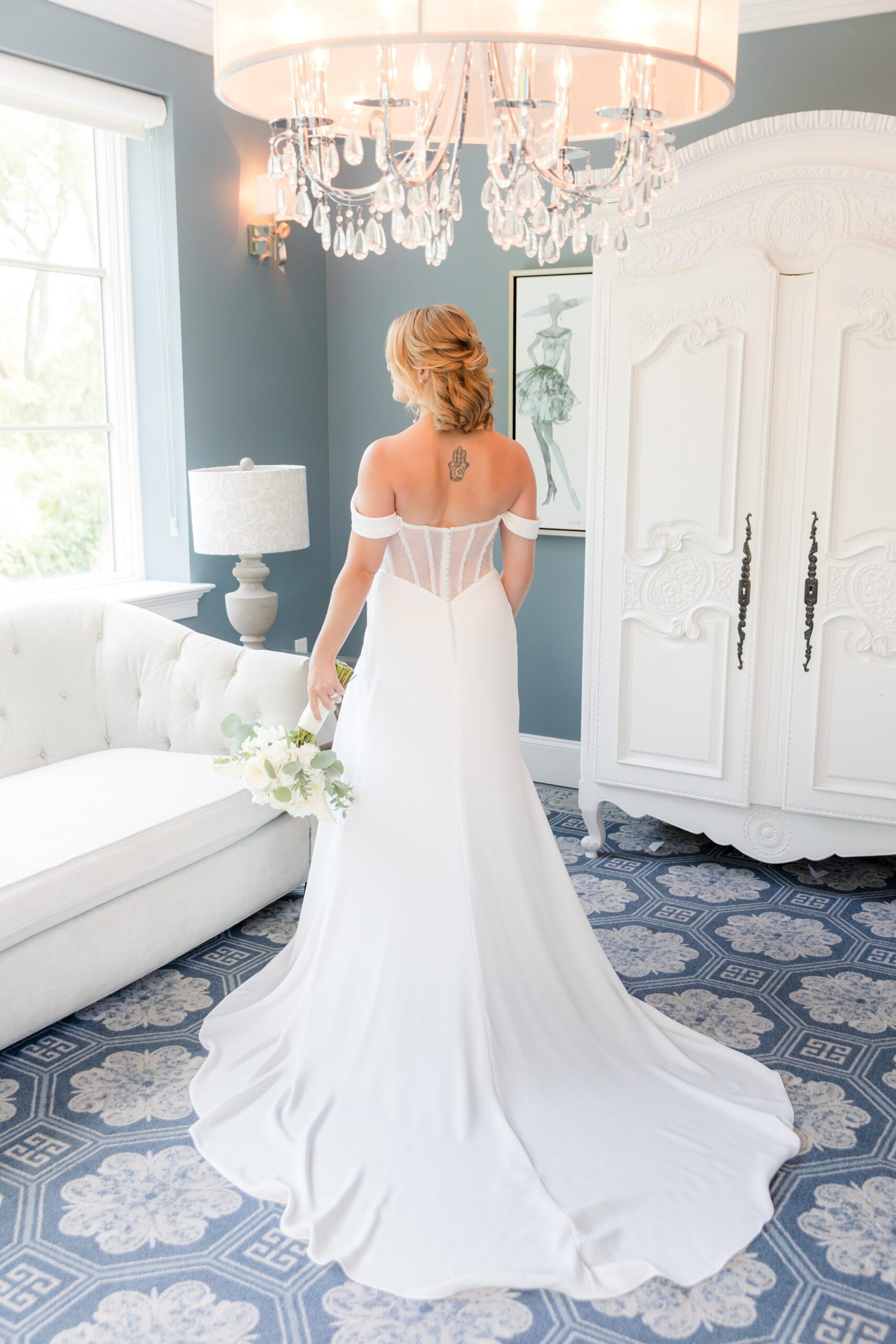 Bride stands with her back turned, her gown cascading behind her in soft, elegant folds beneath a glowing chandelier