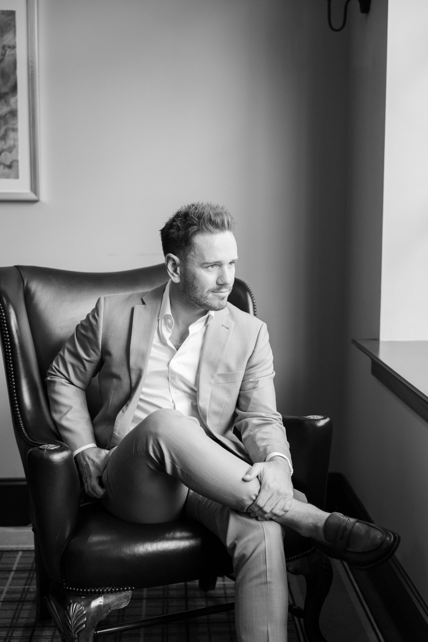 Groom sits quietly by the window, lost in thought as soft light falls across his face, capturing a calm, reflective moment before the celebration begins
