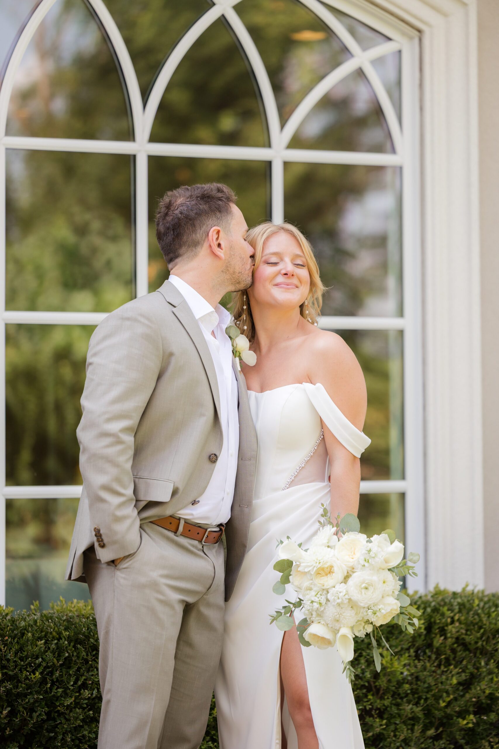 Groom kisses the bride’s cheek as she closes her eyes, smiling softly while holding a bouquet of white flowers