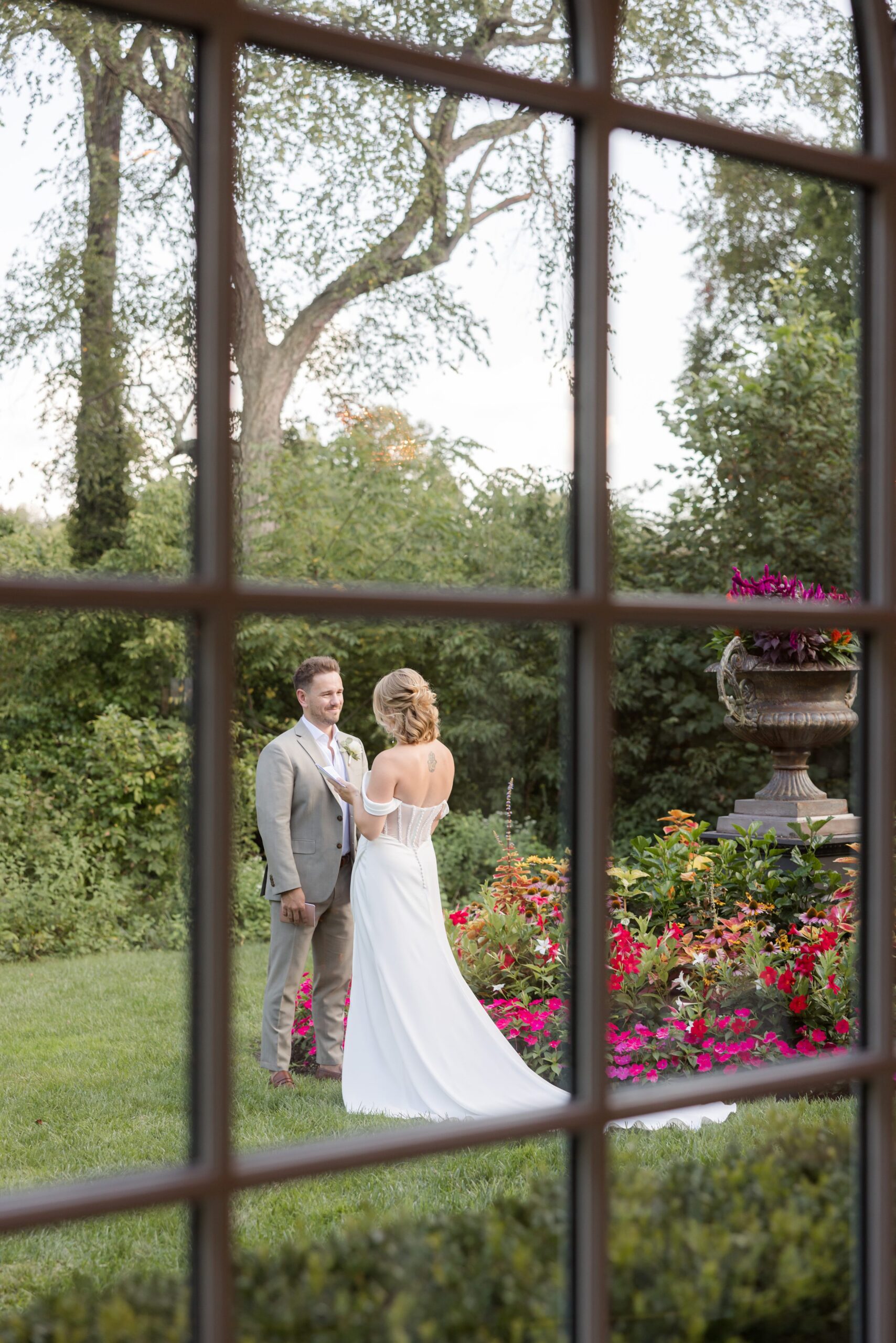Bride and groom stand close together in a quiet, intimate moment, framed by a window and blooming garden beyond