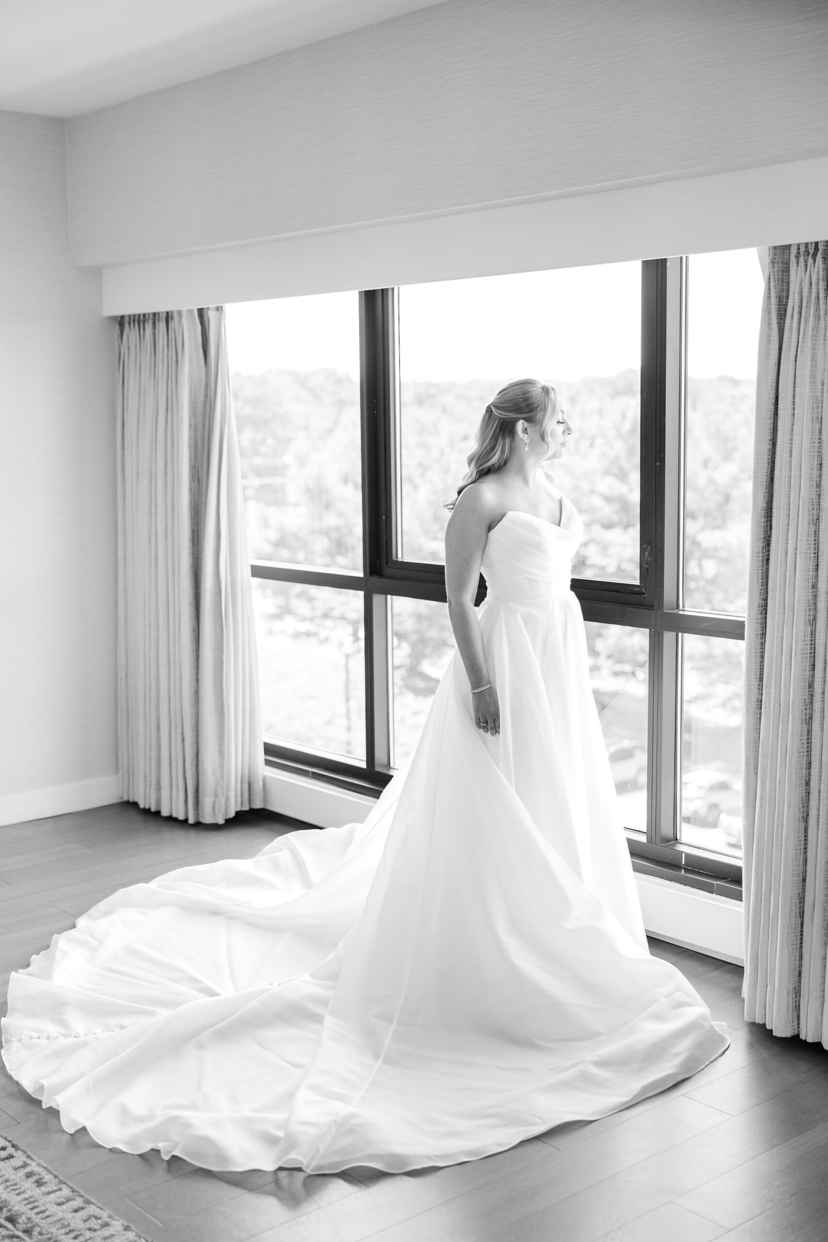 Black-and-white image of a bride standing by a large window, looking outside, her wedding gown’s long train spread across the floor and softly lit by natural light