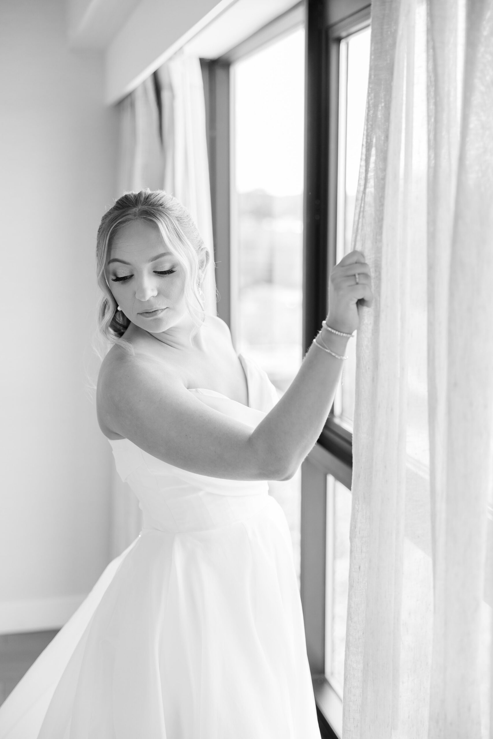 Bride standing by a window, gently holding the curtain and looking down, her strapless wedding gown softly illuminated by natural light
