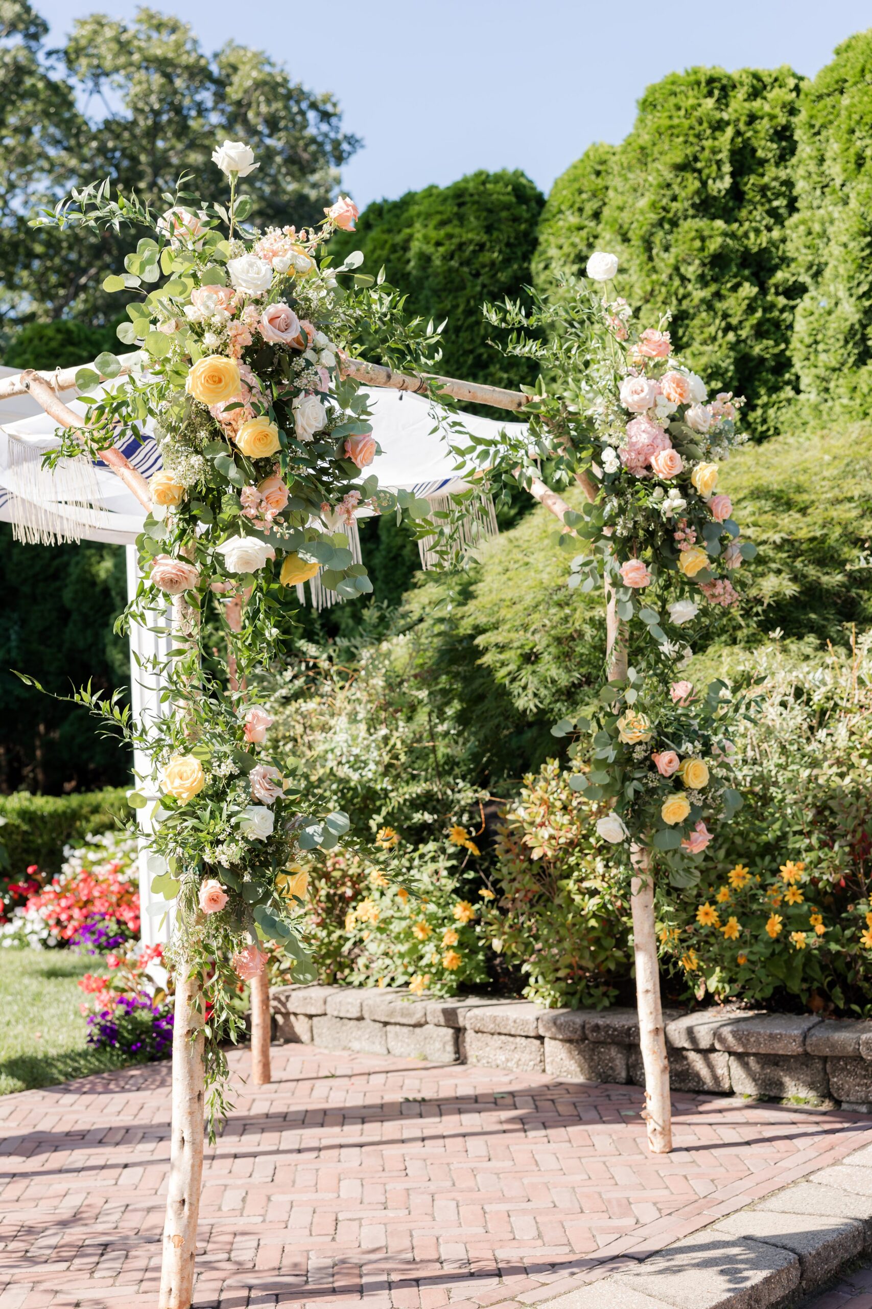 Wedding arch adorned with lush greenery and pastel flowers, set in a garden with vibrant blooms and natural sunlight creating a romantic ceremony backdrop