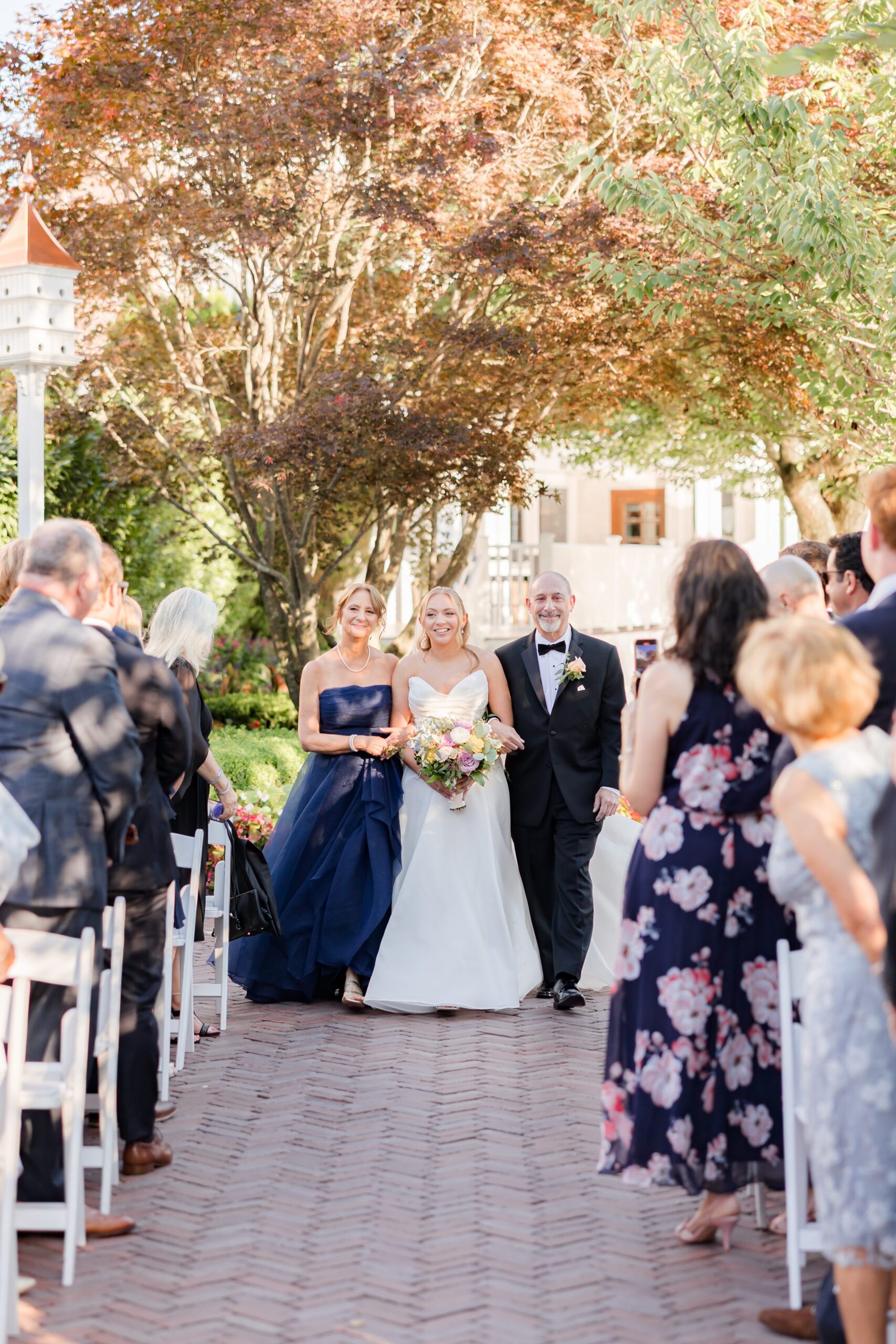 Bride walking down the aisle with her parents during an outdoor ceremony, smiling as guests stand on either side, surrounded by soft sunlight and greenery