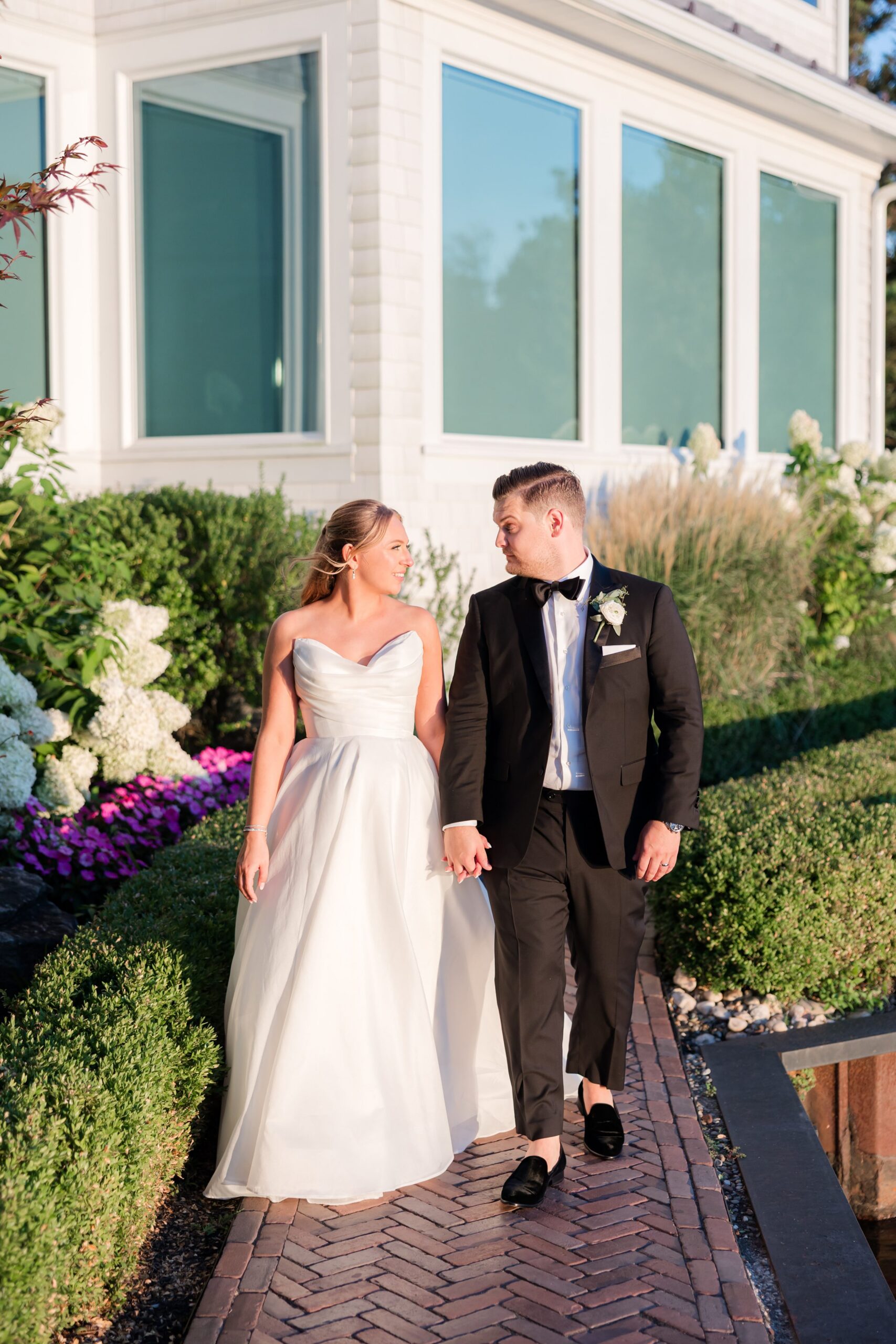 Bride and groom walking together along a garden path, smiling at each other in warm sunlight, surrounded by greenery and blooming flowers in a romantic moment