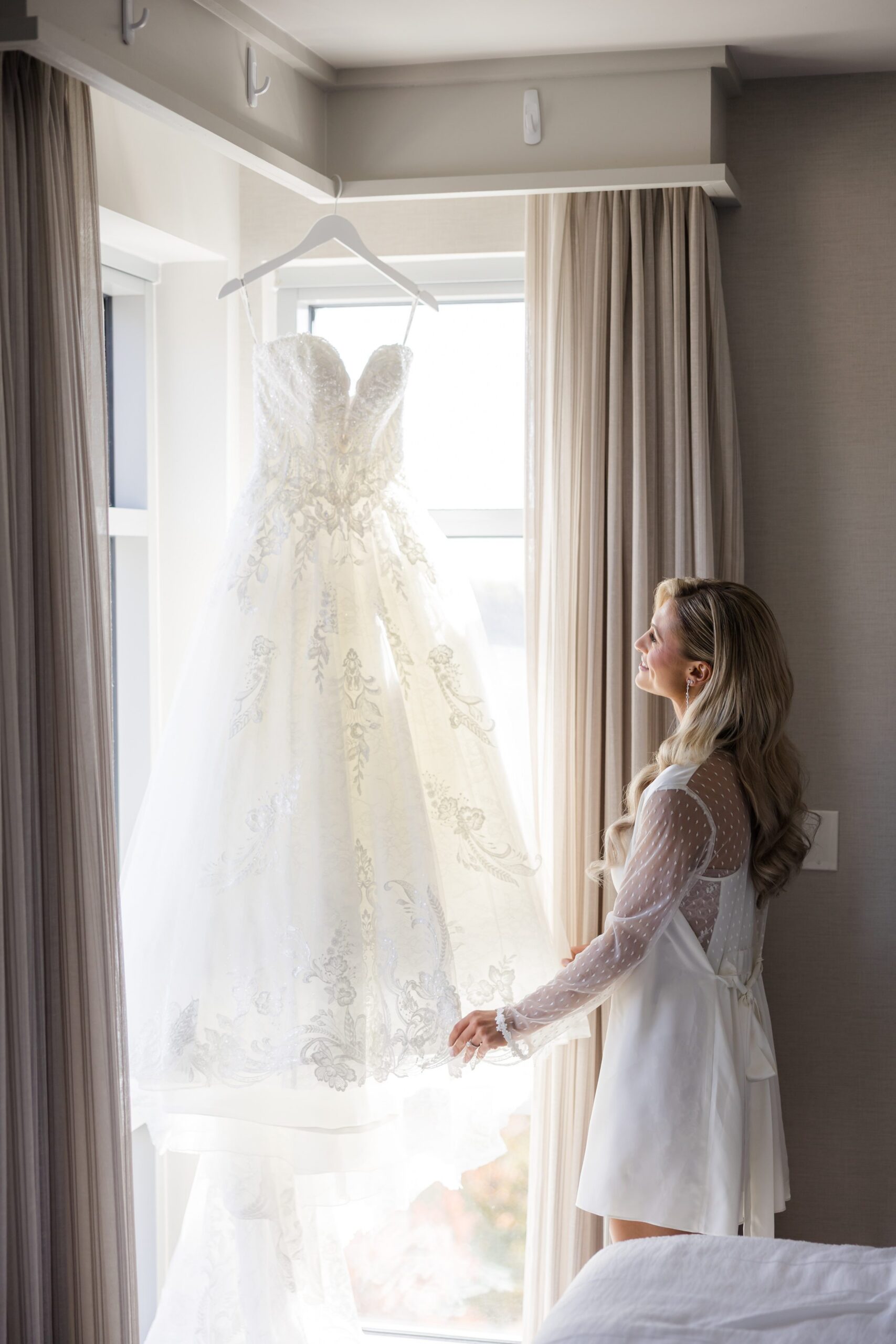 Bride in a robe standing by a window, admiring her lace wedding dress hanging in soft natural light, framed by sheer curtains