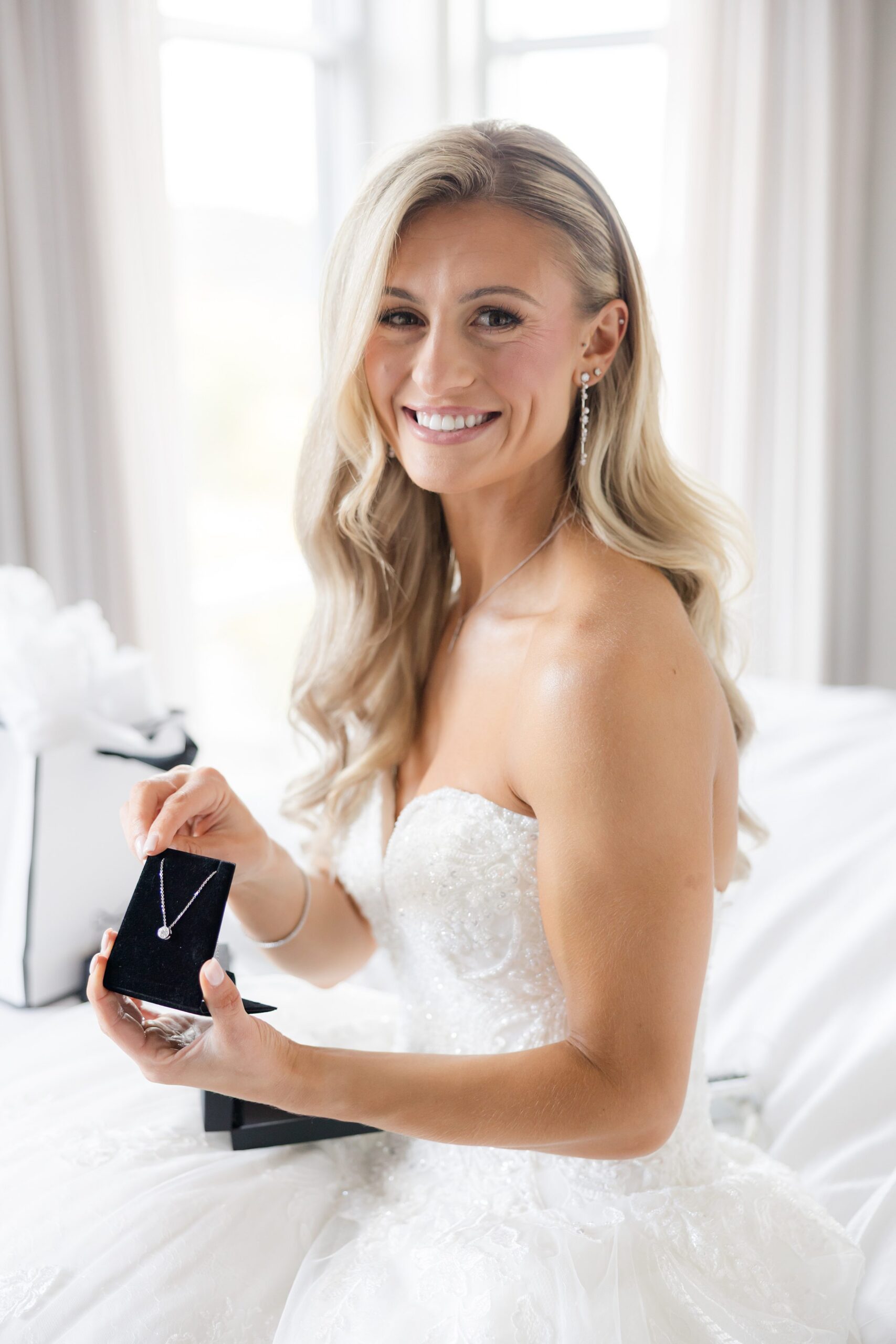 Bride in a white strapless wedding dress sits on a bed in a bright room, holding an open jewelry box with a necklace inside
