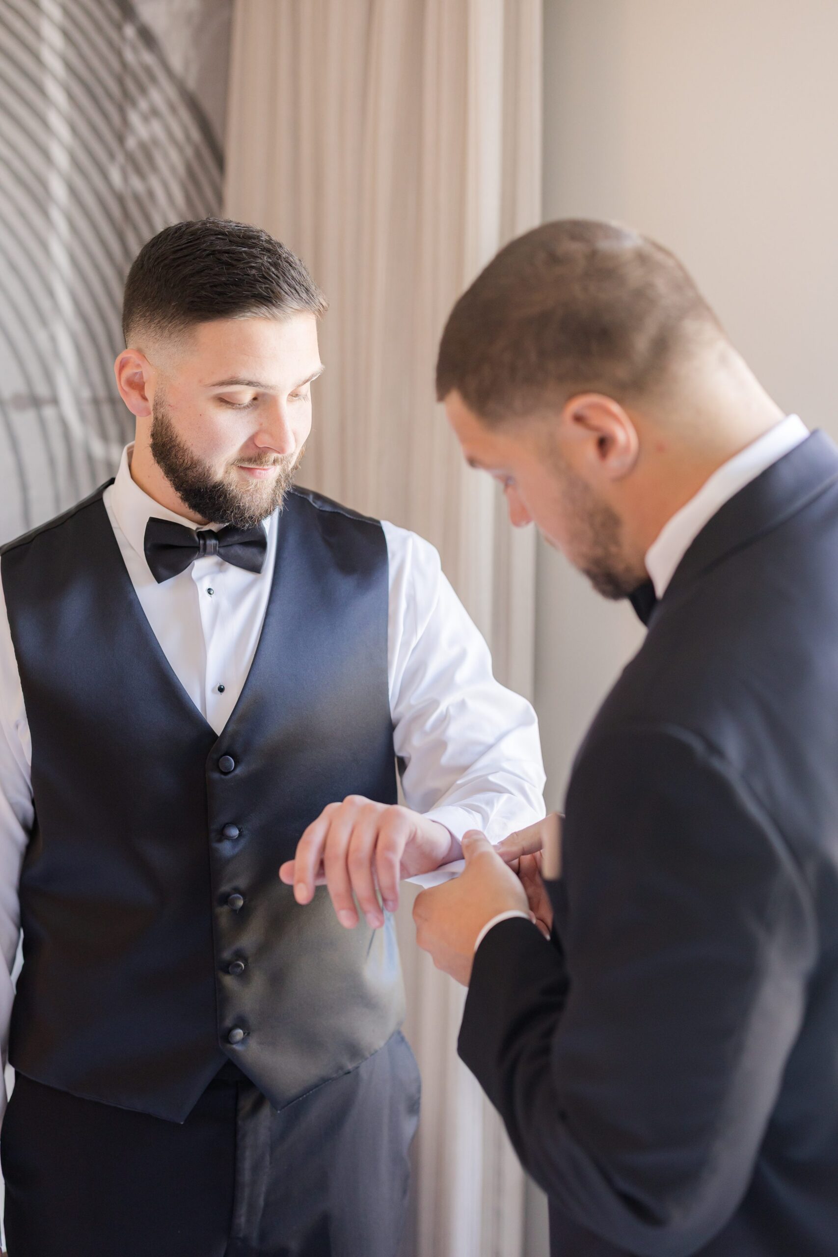 Groom and groomsman sharing a quiet moment while getting ready, adjusting cufflinks and final details, both dressed in formal suits by a window with soft natural light