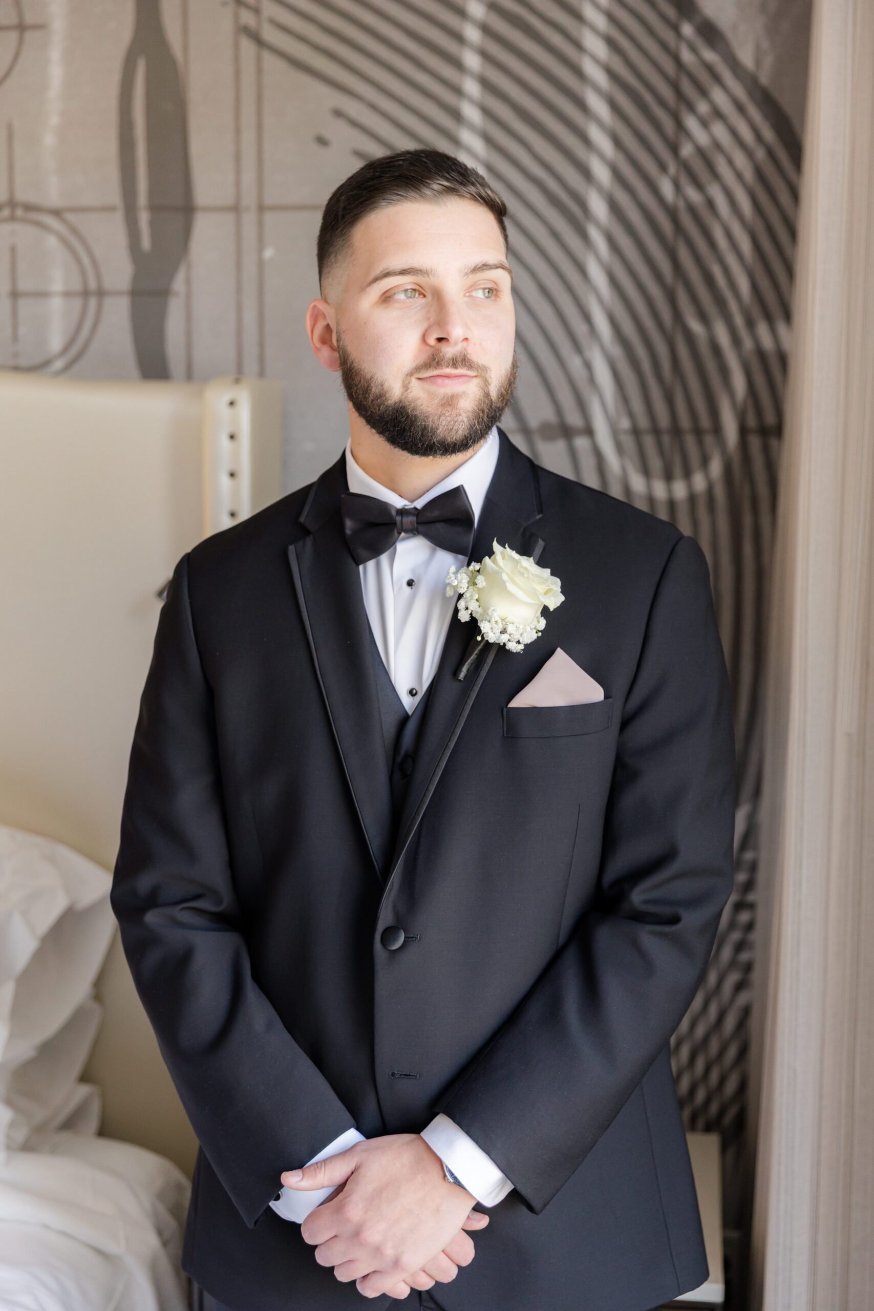 Groom in a classic black tuxedo with bow tie, white boutonniere, and pocket square, standing by a window with hands clasped, softly lit as he looks off to the side in a calm, reflective moment