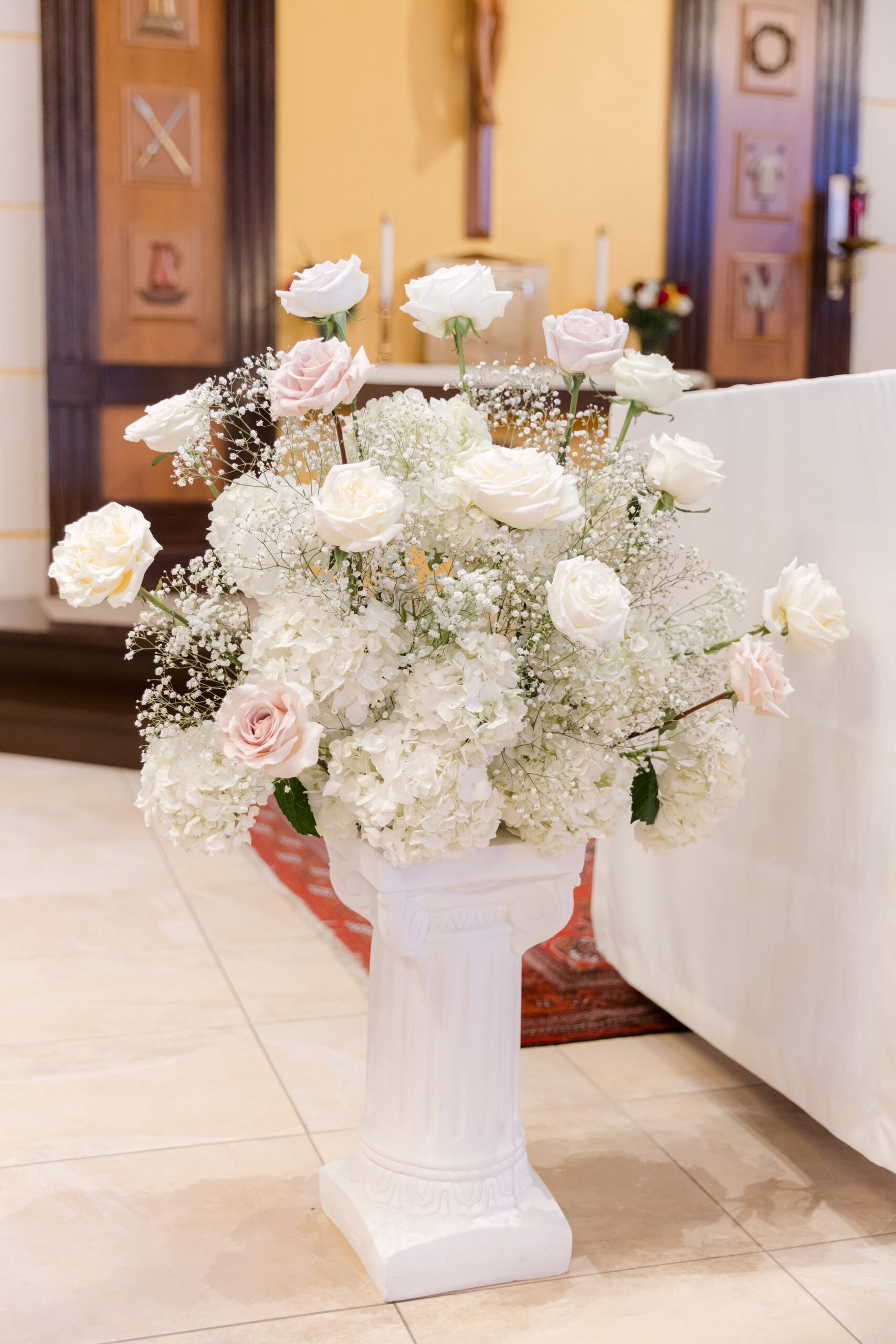 Elegant white floral arrangement of roses and baby’s breath placed on a pedestal inside a church, softly lit and positioned near the altar for a wedding ceremony