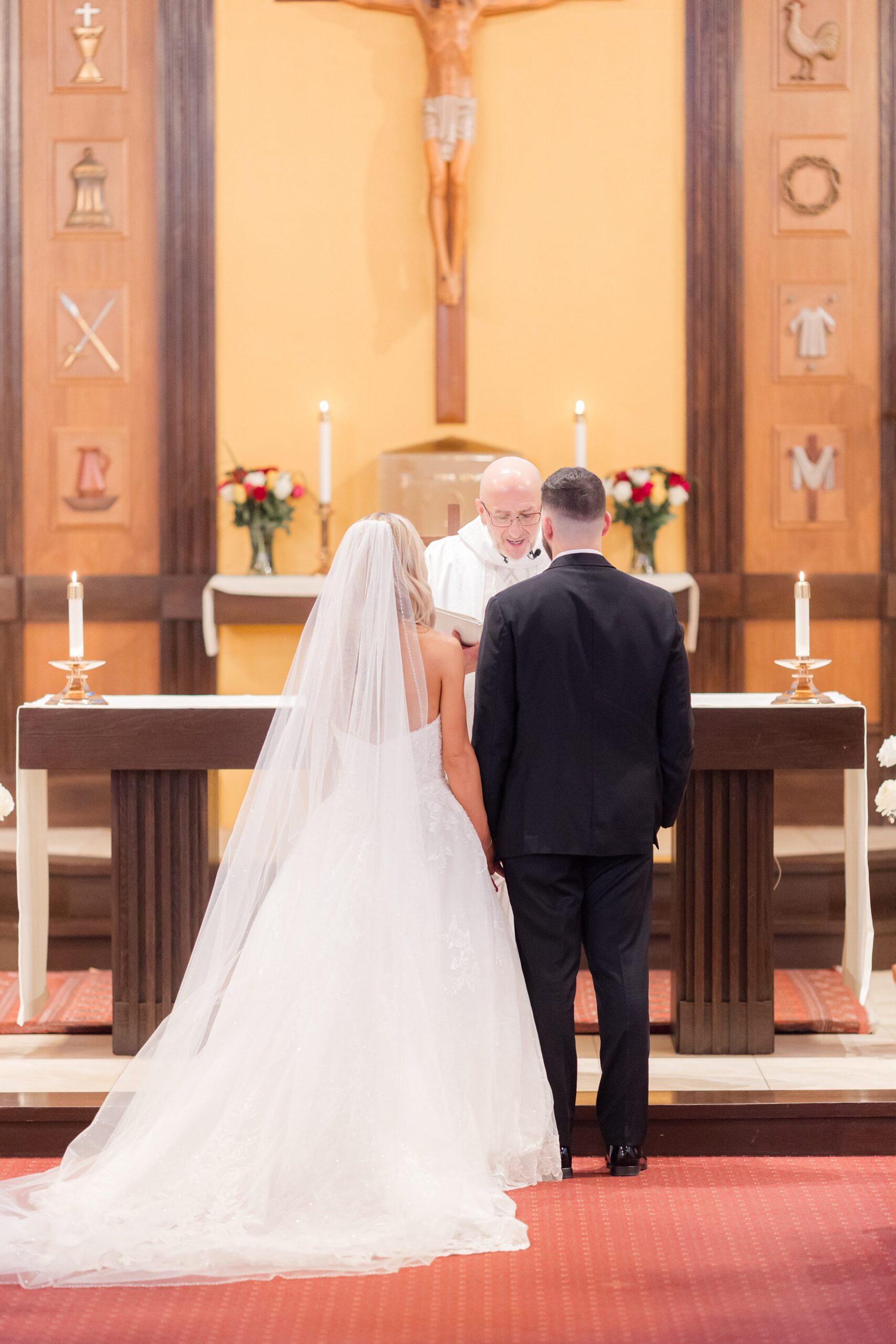 Bride and groom standing side by side at the altar during their church wedding ceremony, facing the officiant beneath a large crucifix, with candles and floral arrangements creating a solemn, romantic setting