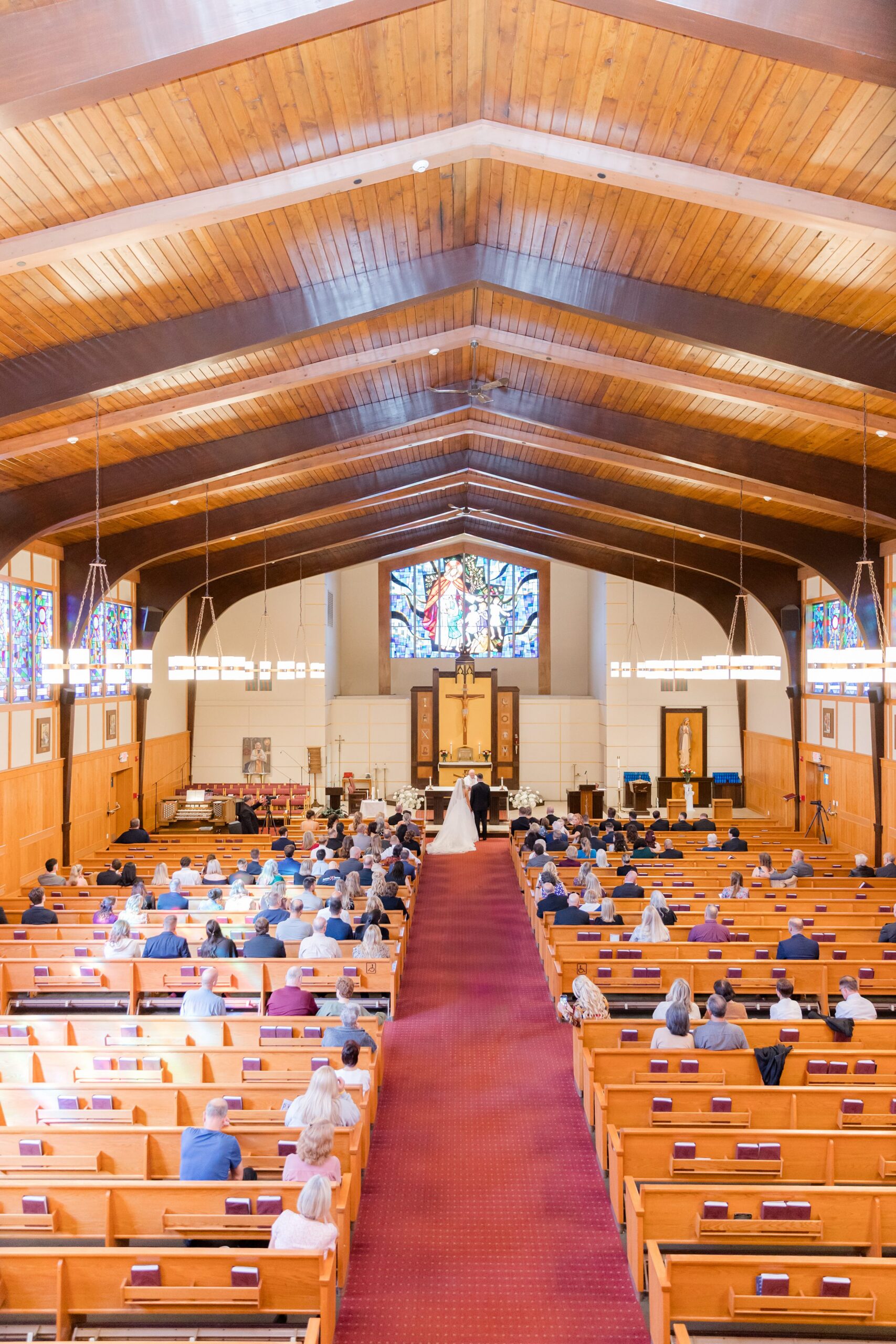 Wide view of a church wedding ceremony with the bride and groom at the altar, guests seated in wooden pews along a red aisle, and a stained glass window above