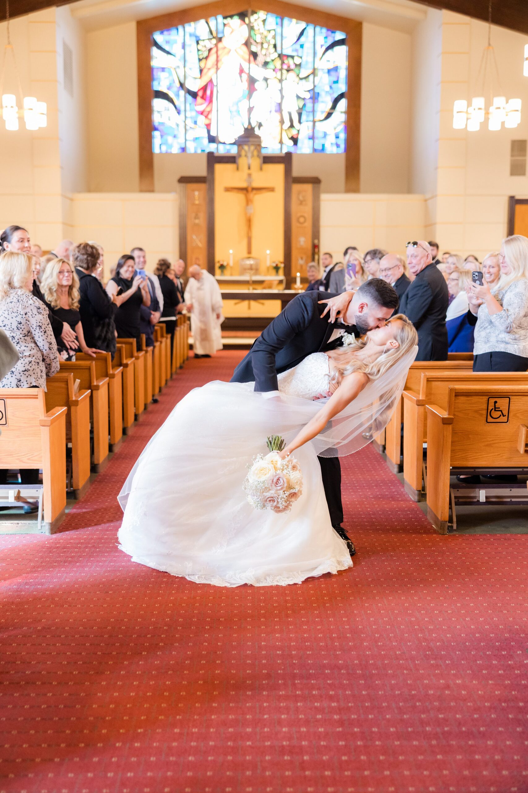 Groom dipping the bride for a kiss in the church aisle as guests stand and watch, celebrating their joyful wedding moment