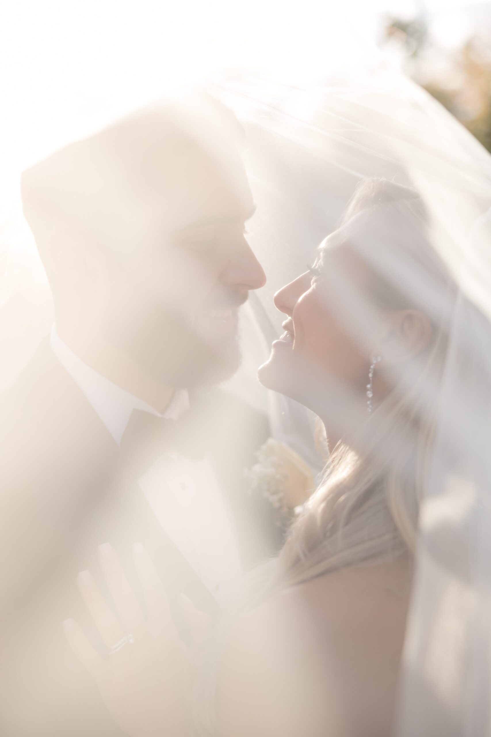 Bride and groom gazing at each other beneath the veil, softly backlit to create a dreamy, romantic wedding moment
