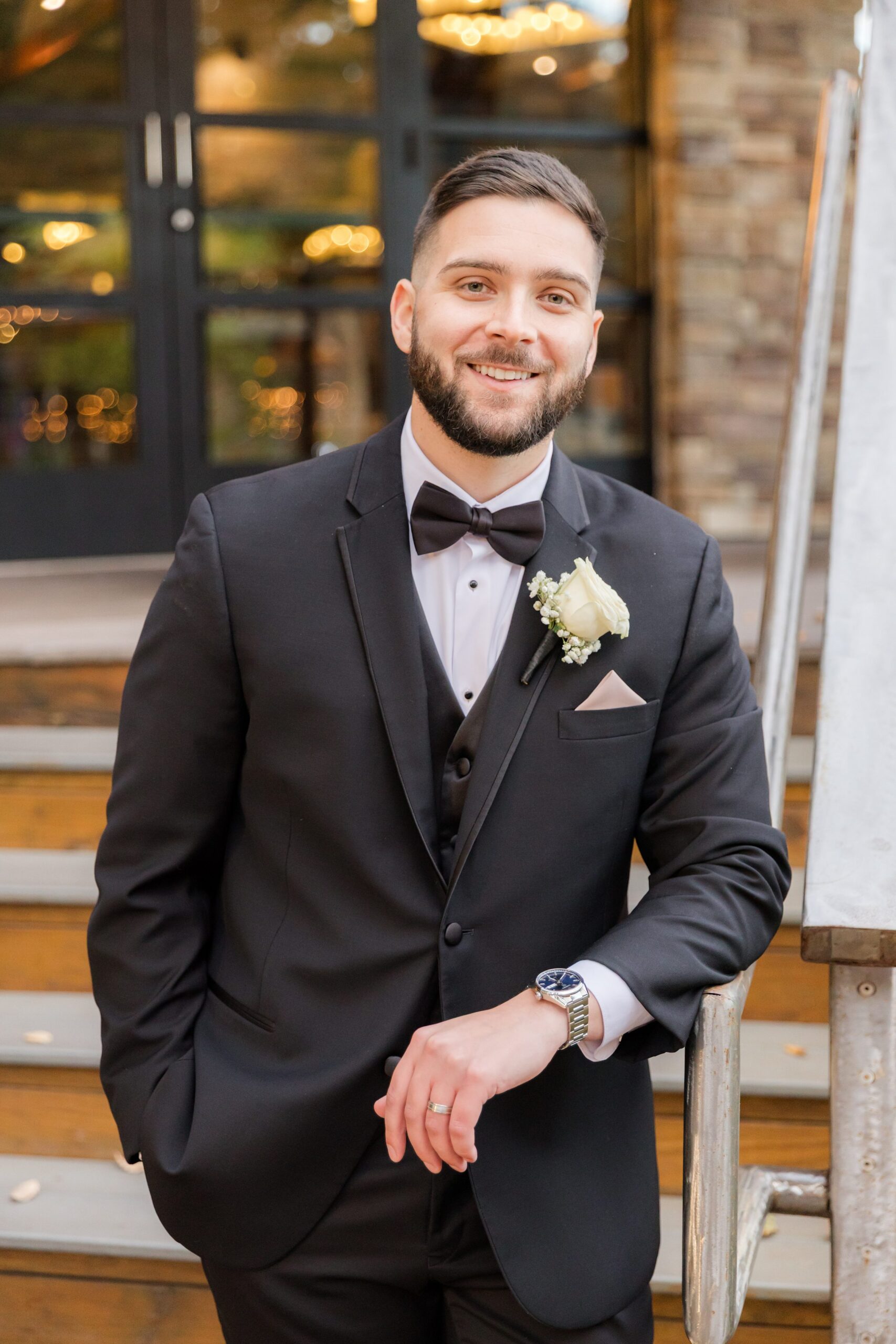 Groom in a black tuxedo with a bow tie and boutonniere, smiling while leaning on a railing outside, captured in a relaxed and confident portrait