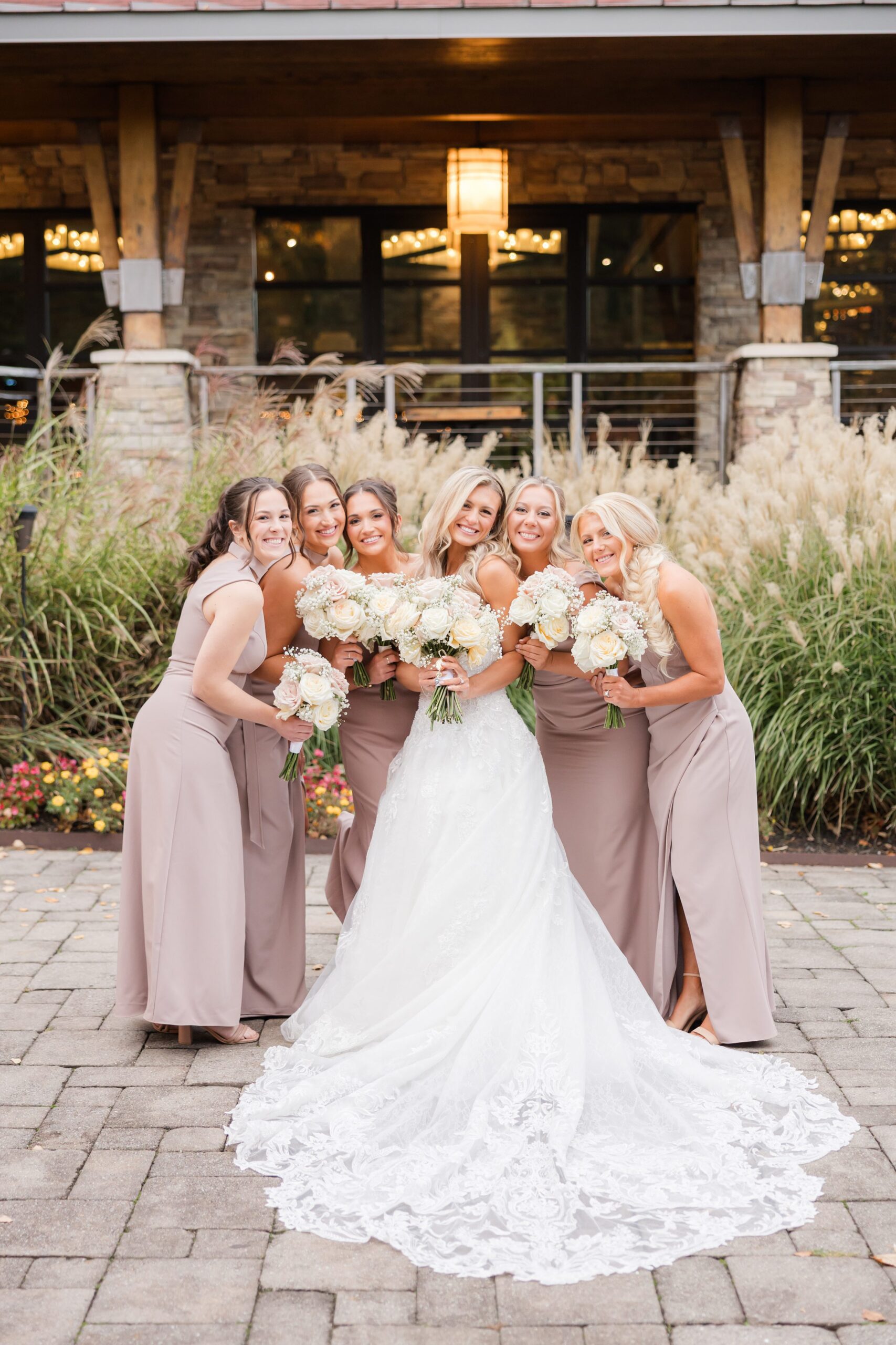 Bride standing with bridesmaids in matching dresses, all holding bouquets and smiling together in an outdoor wedding portrait