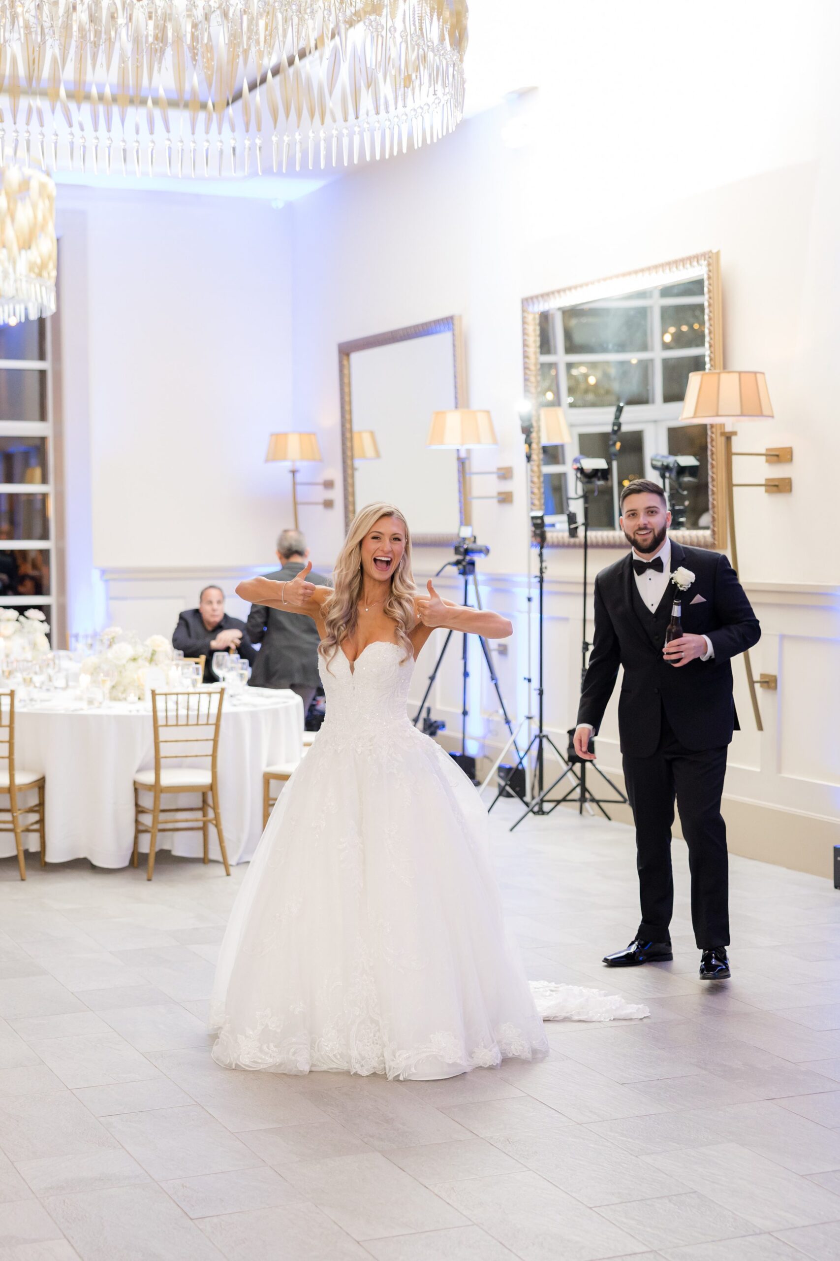 Bride and groom smiling and dancing in an elegant, well-lit wedding reception hall with chandeliers