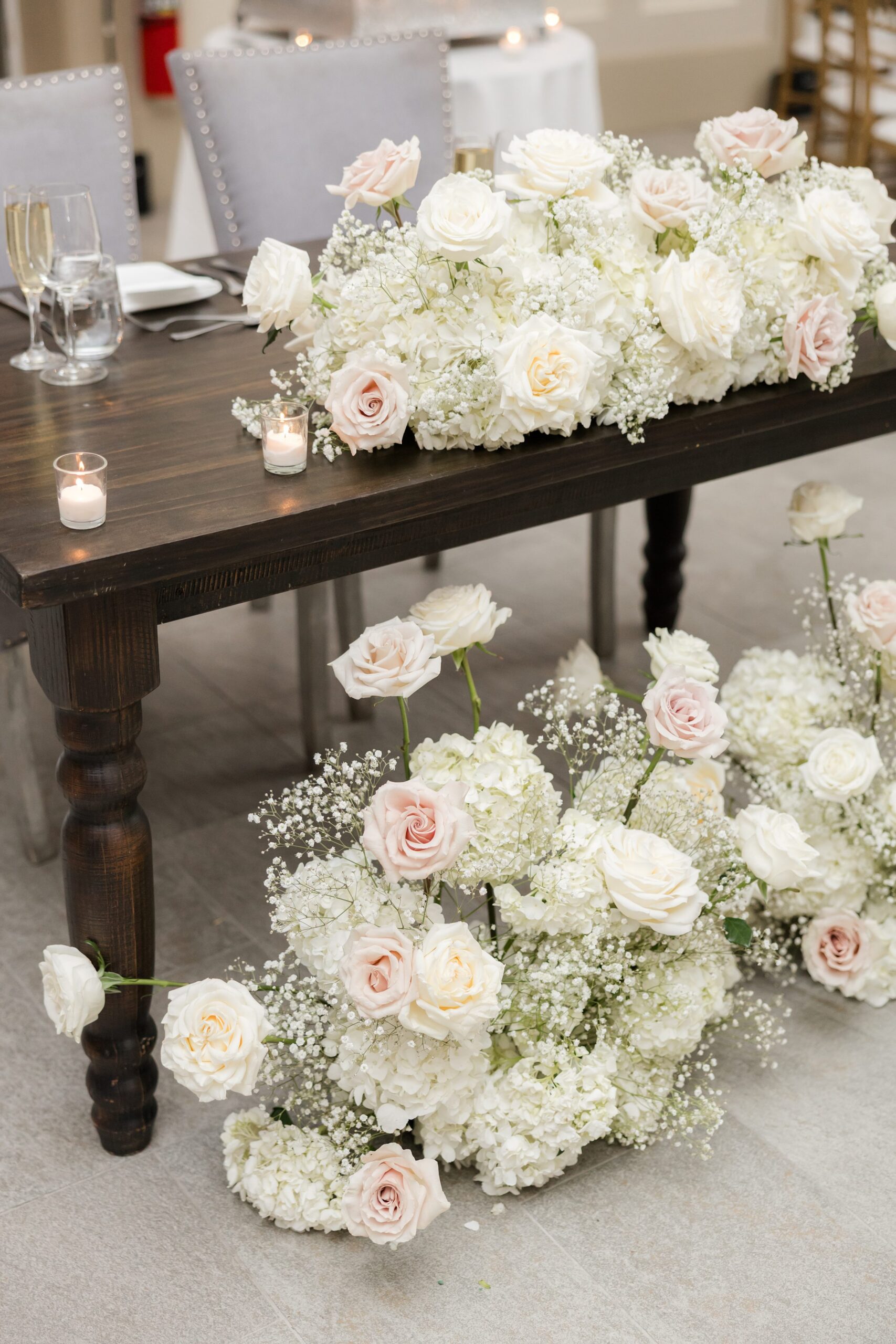 Dark wooden table decorated with white and blush floral arrangements on top and beneath, with small candles and glassware