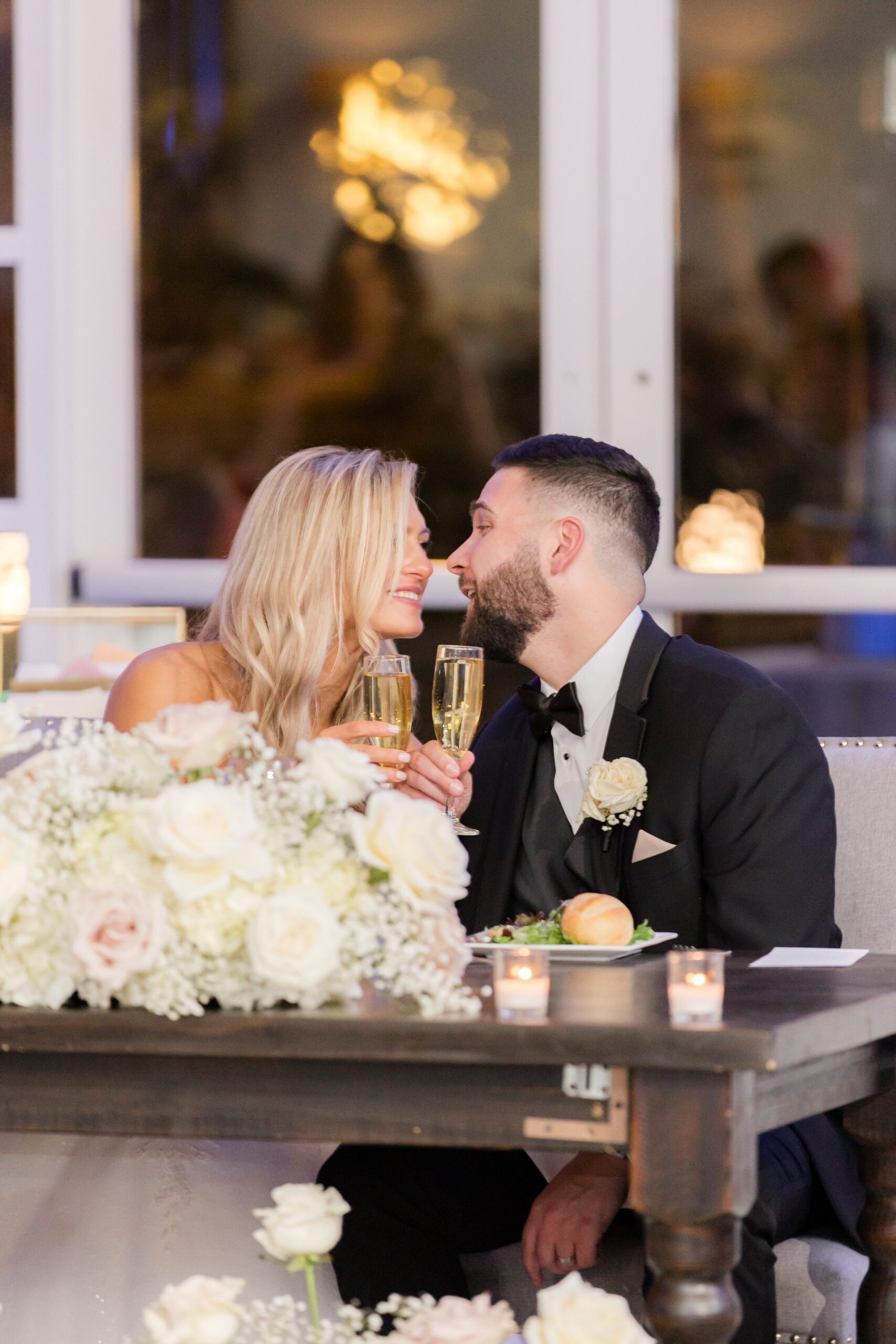 Bride and groom smiling at each other while seated at a decorated table with flowers and champagne