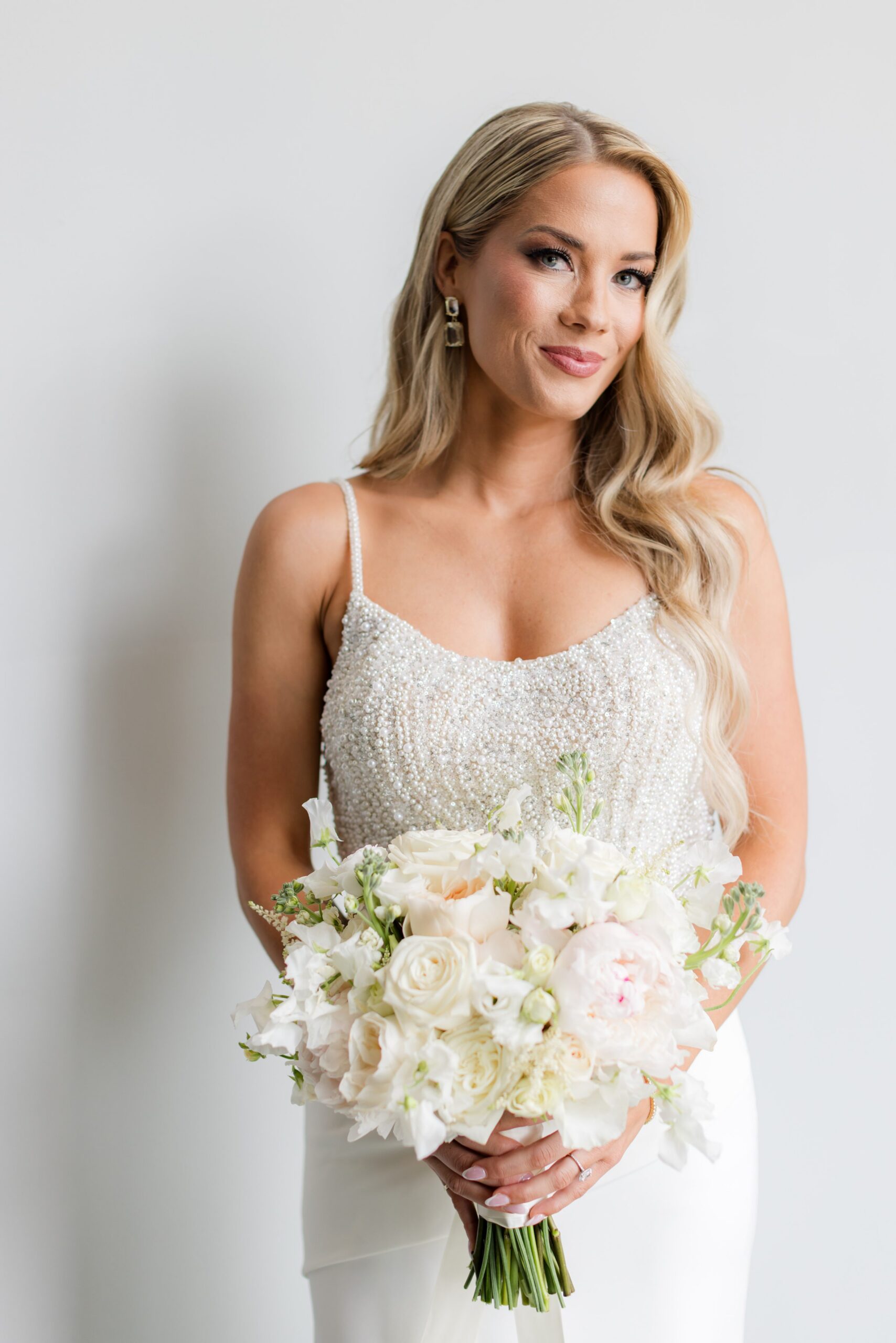 Bride in a beaded wedding gown holding a bouquet of white flowers, posing softly against a neutral background