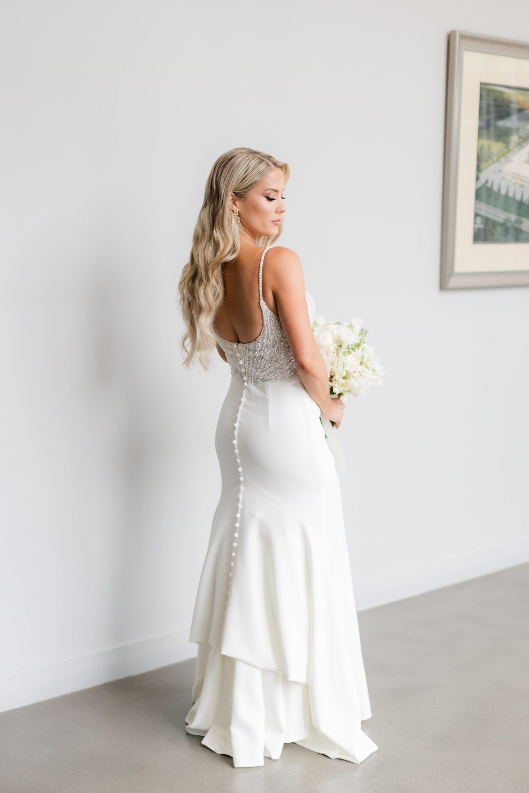 Bride in a fitted wedding gown viewed from behind, holding a bouquet and showcasing elegant button details along the dress