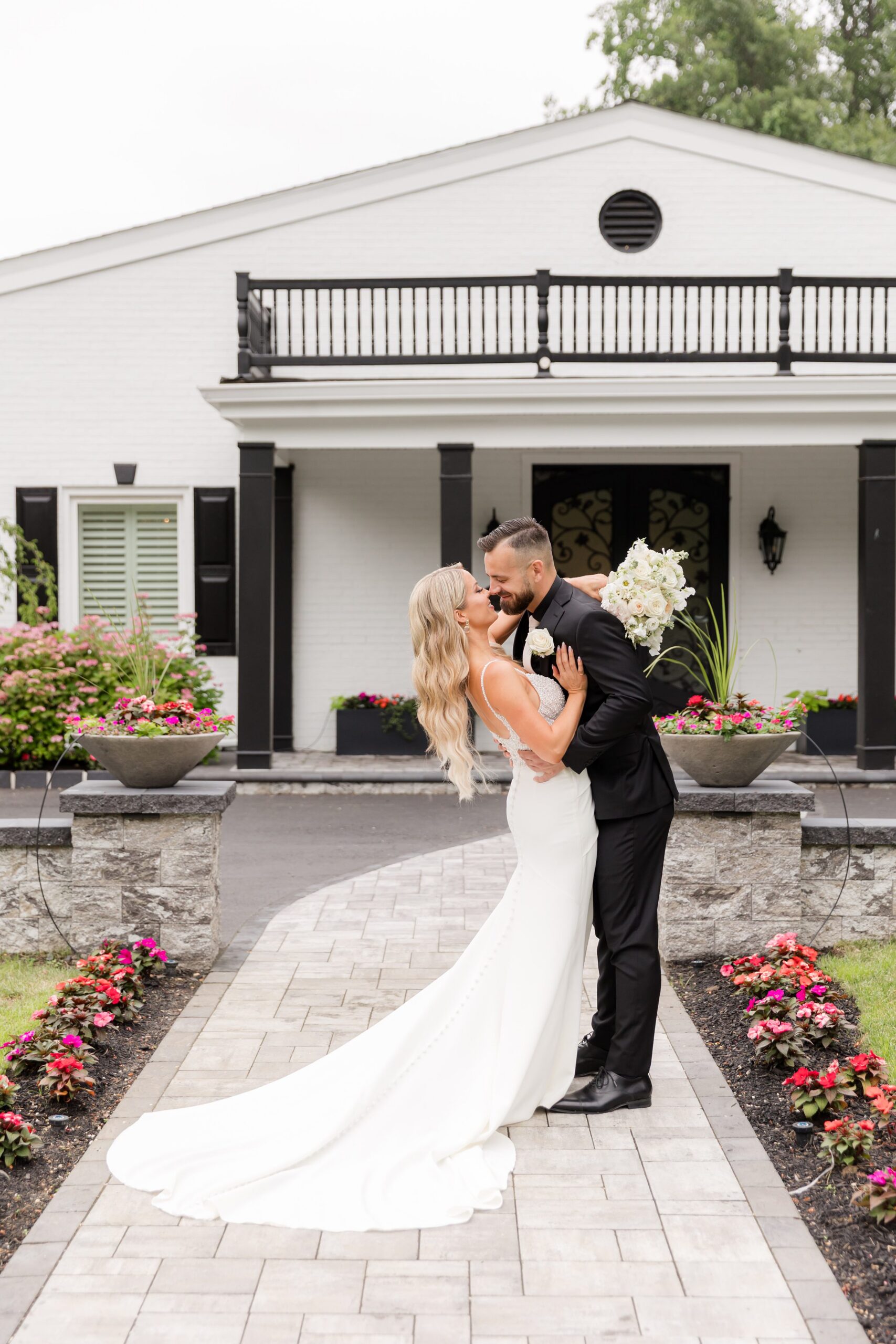 Bride and groom sharing a romantic dip and kiss on a garden path in front of their venue, with her gown flowing elegantly behind her
