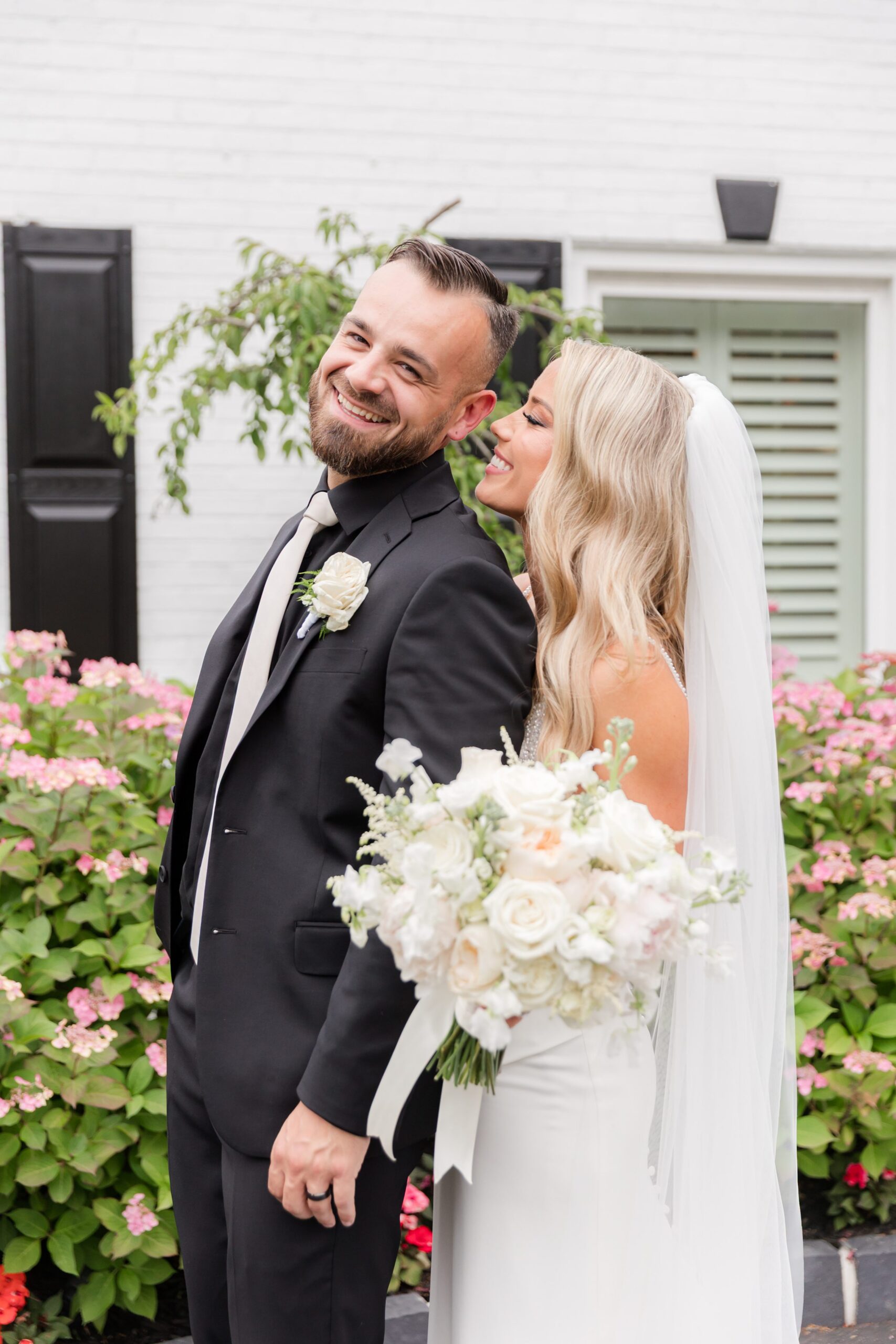 Bride leaning close to the groom as he smiles, holding her bouquet and sharing a playful, affectionate moment outside the venue