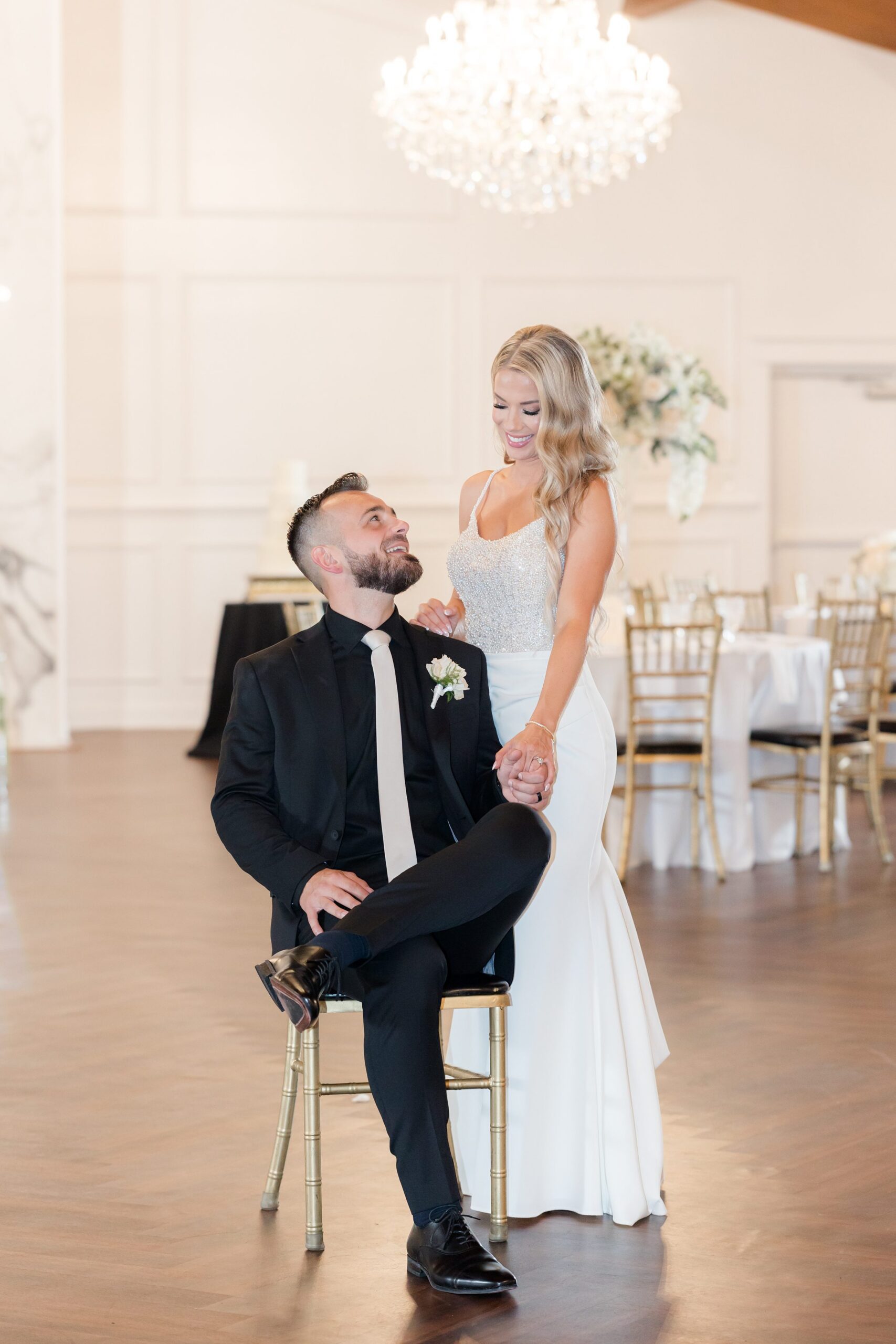 Bride and groom sharing a playful reception moment, with the groom seated and looking up as the bride smiles beside him under elegant chandeliers