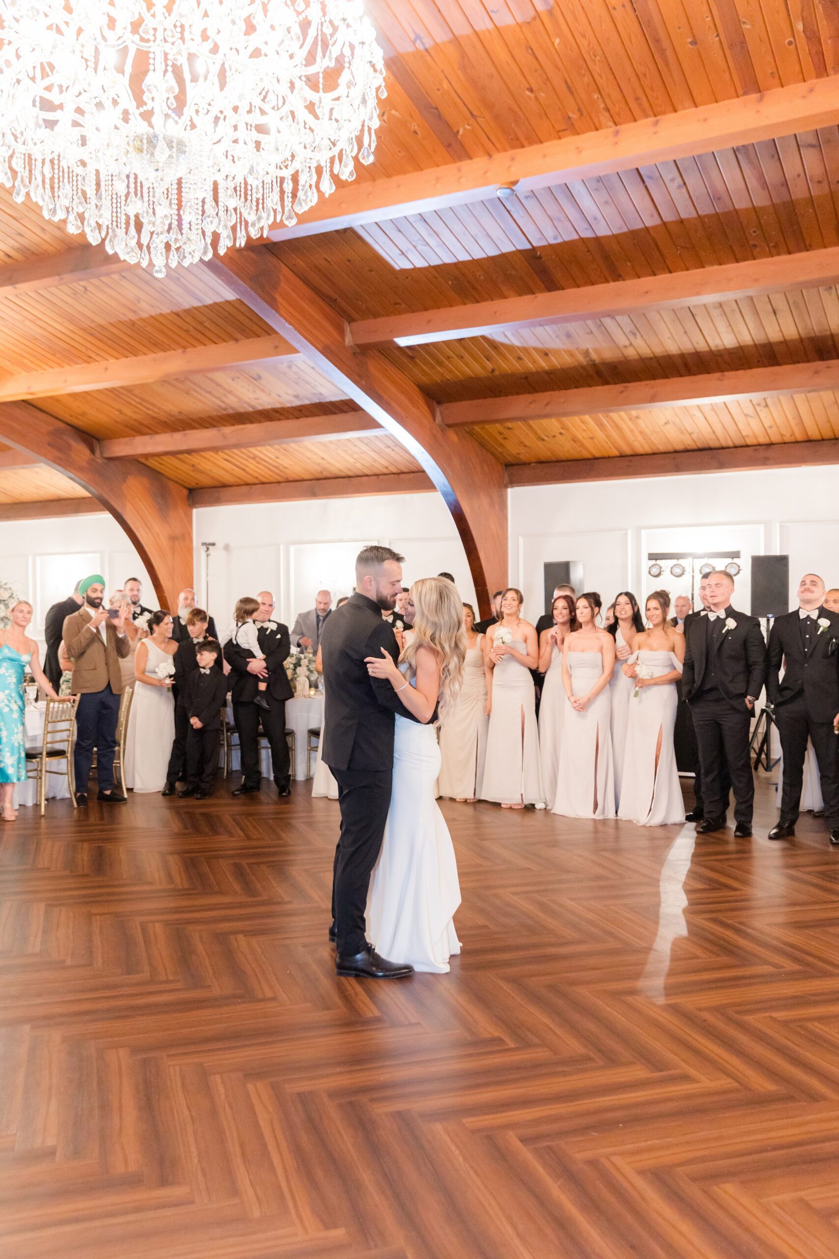 Bride and groom sharing a romantic first dance in the center of the reception floor, surrounded by their wedding party and guests watching