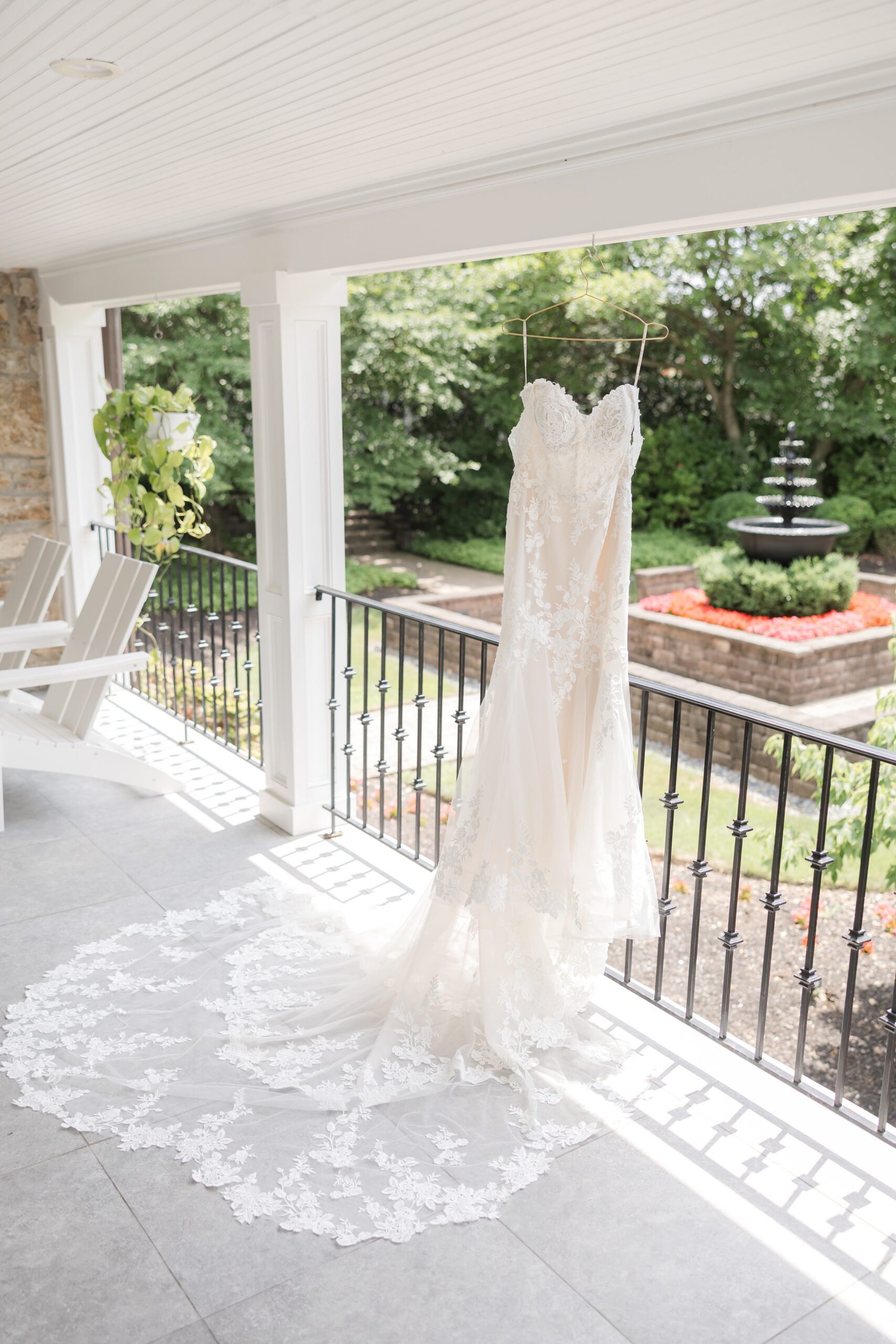 Wedding dress hanging on a balcony railing, with a long lace train flowing down and a garden fountain in the background