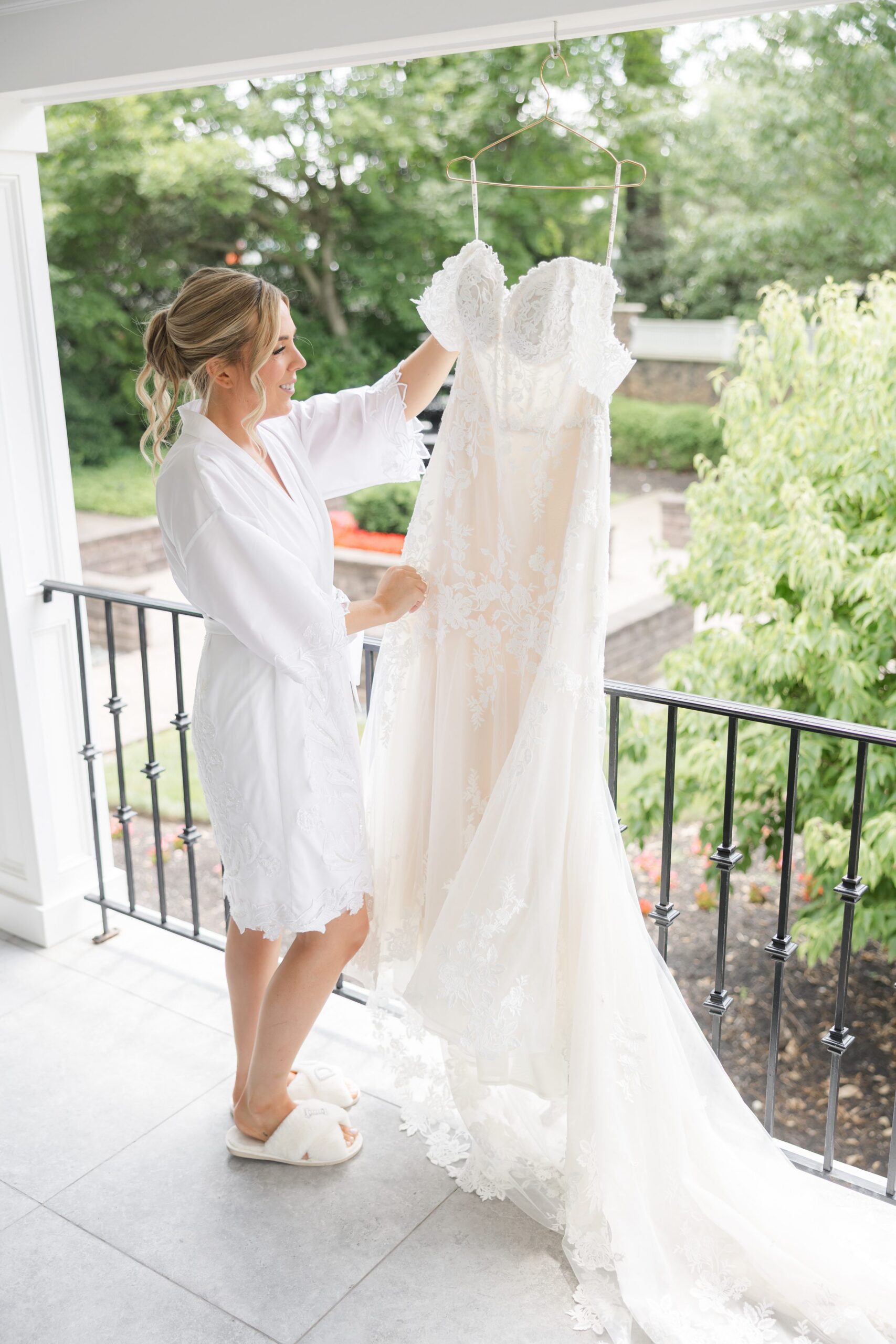Bride in a robe admiring her wedding dress hanging on a balcony, with the gown’s lace train flowing and greenery in the background