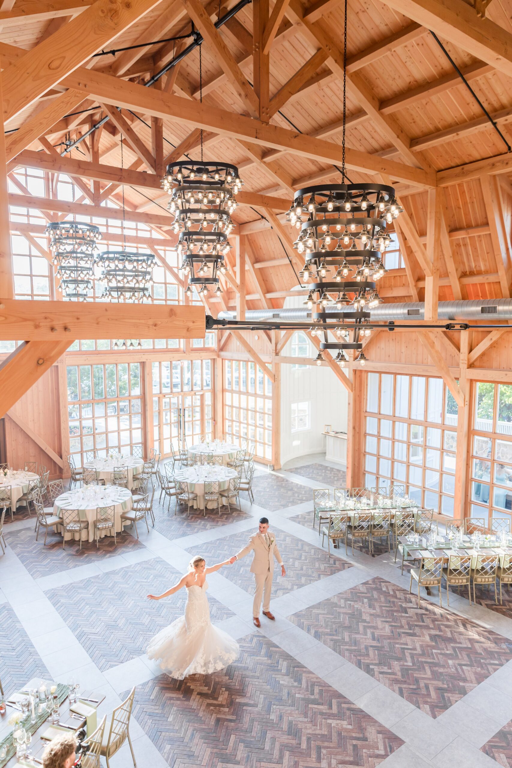 Bride and groom dancing together in the center of a large, sunlit reception hall with high wooden ceilings, grand chandeliers, and tables arranged around them