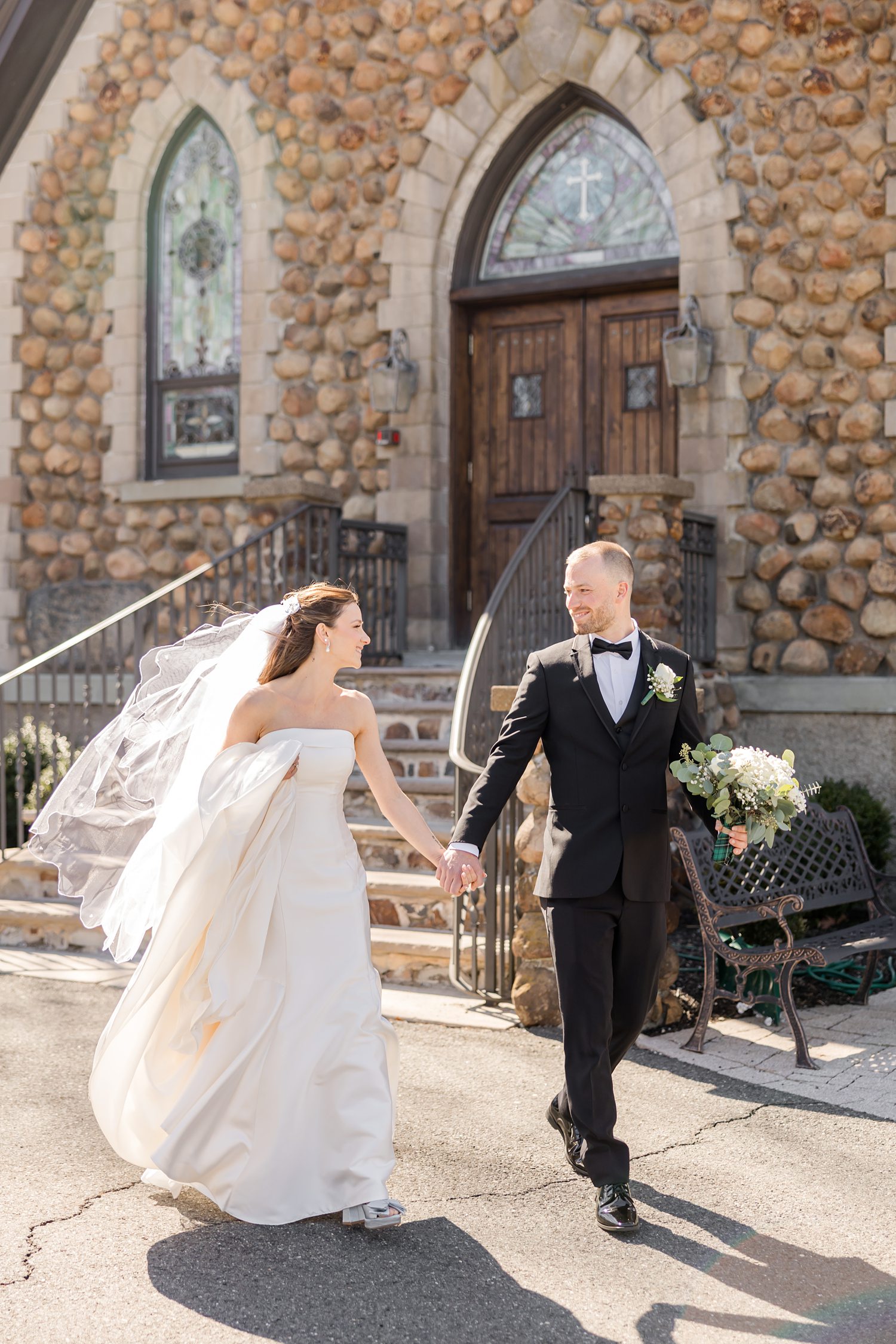 Bride and groom walking outside church with veil flowing in spring sunlight